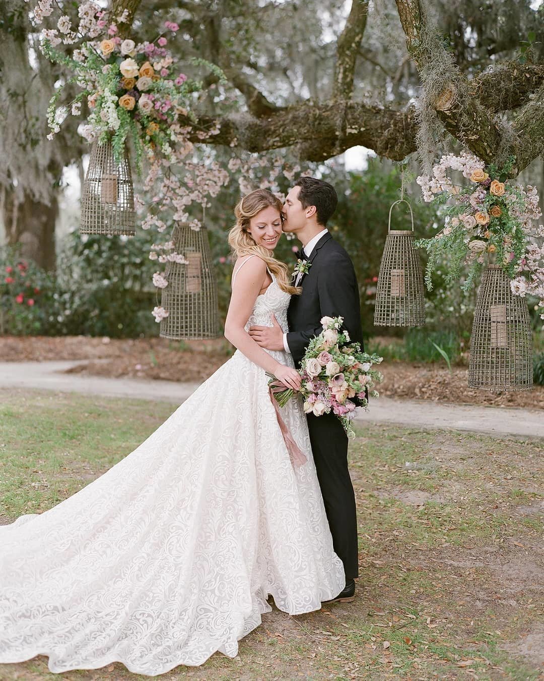 Live oak tree wedding backdrop with floral adorned rattan pendants and a fashionable bride and groom