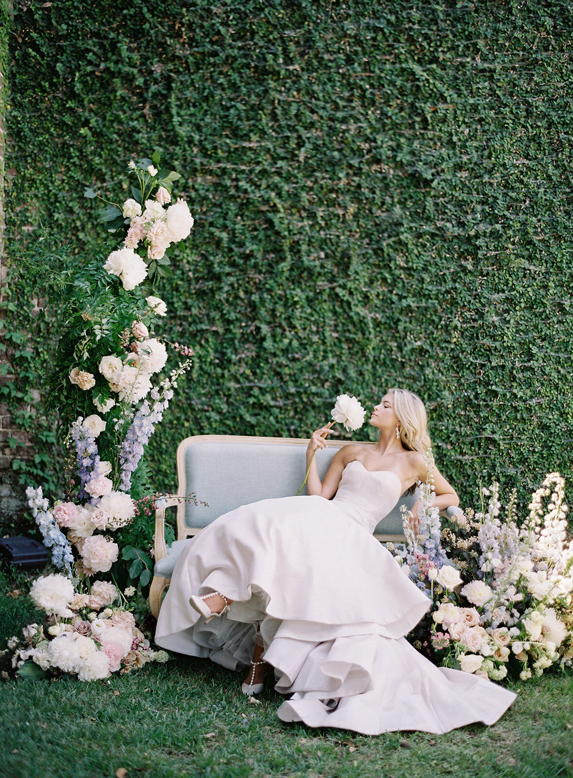 Middleton Place Wedding. Bride on a vintage blue couch in front of a wall of ivy, florals all around her. Bride is in a strapless ivory wedding gown with fitted bodice and flowing train designed by  Anne Barge. Bride has legs crossed and her Bella Belle Shoes ivory heels with pearls showing. She is leaning back holding a delilah flower and smelling it. Photographed by wedding photographers in Charleston Amy Mulder Photography