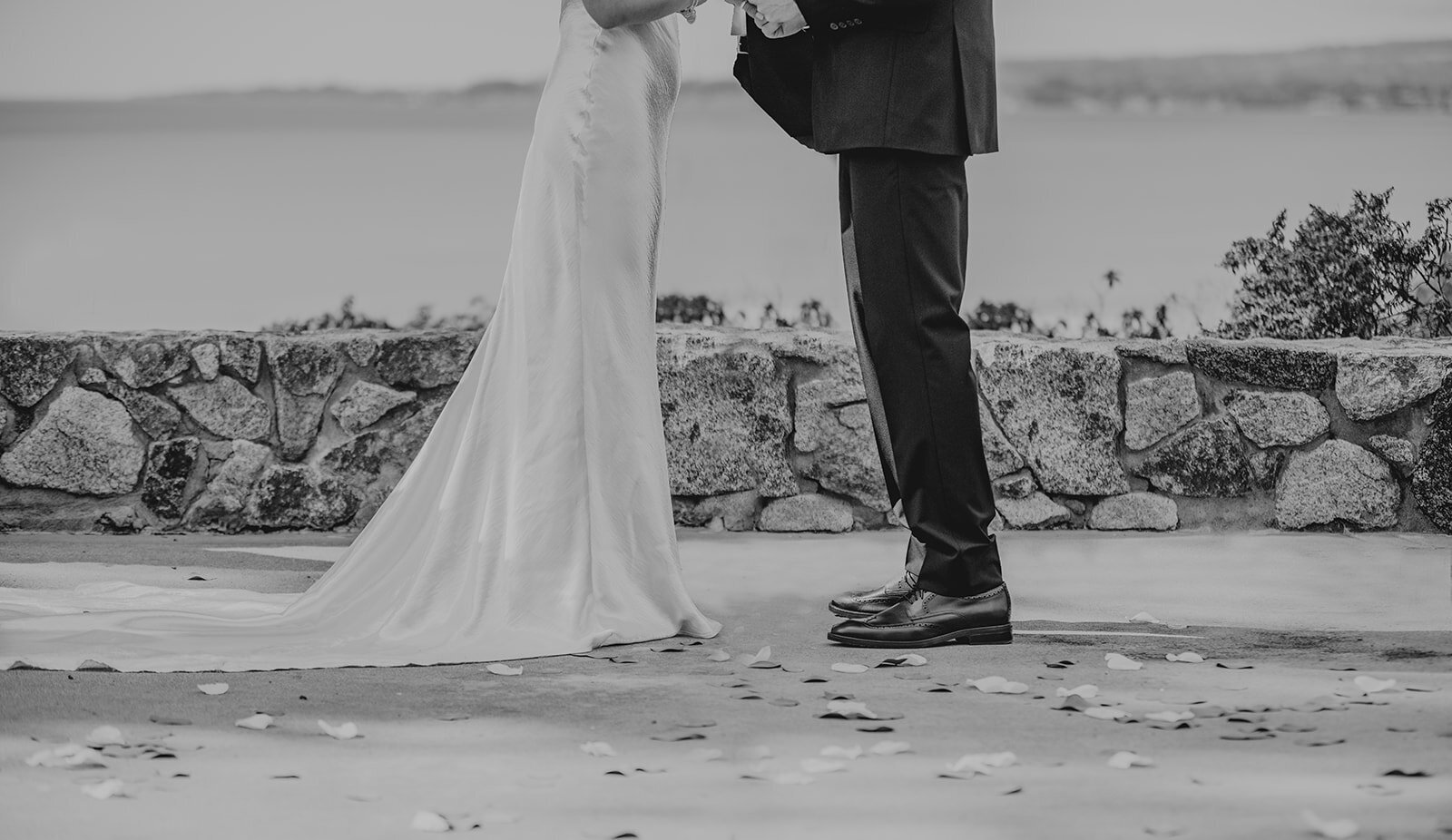 Bride and groom hold hands in an intimate moment by the ocean, with a flowing train and stone wall framing the romantic scene