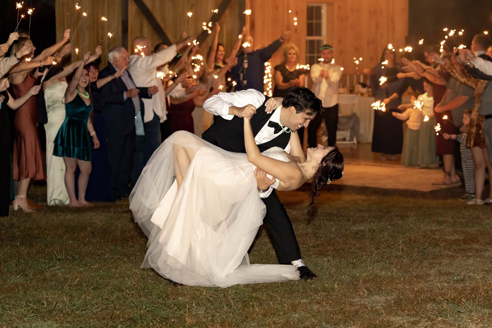 The bride and groom share a romantic dip during an outdoor dance surrounded by guests with sparklers, captured at a rustic barn wedding in Boone, NC, blending celebration and elegance.