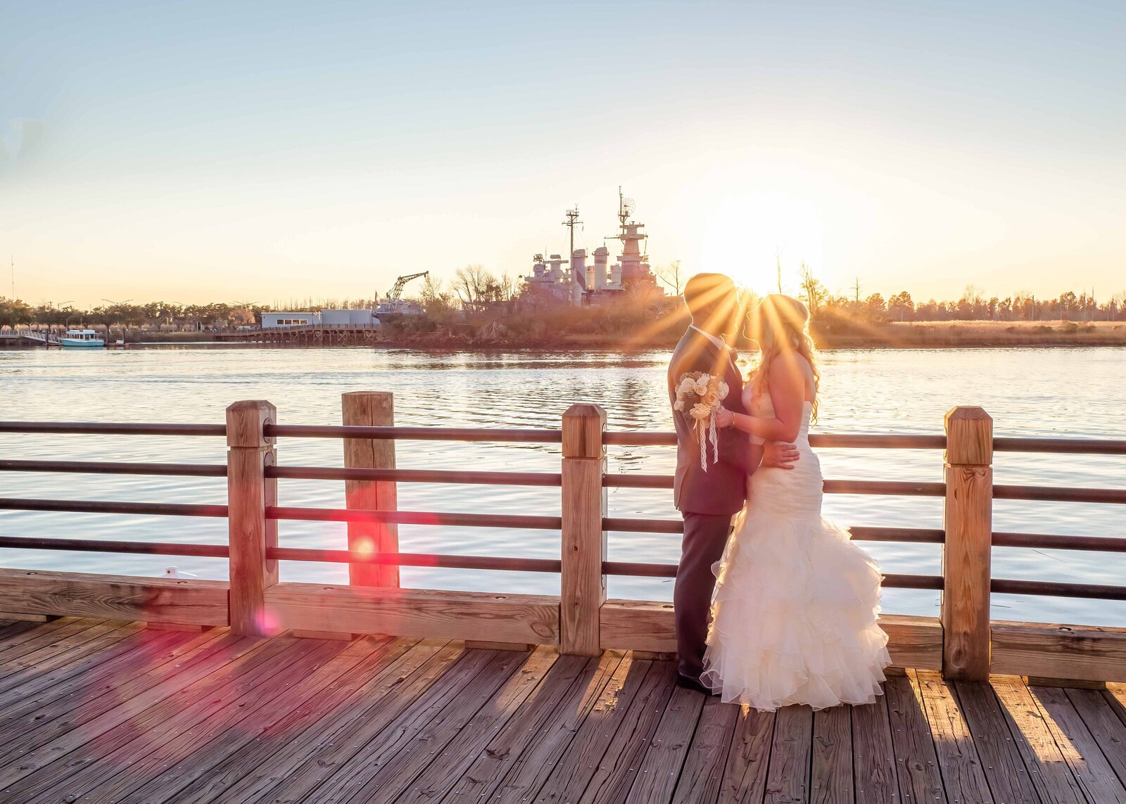The bride and groom share a romantic moment on a boardwalk at sunset with the USS North Carolina Battleship in the background, captured during a Wilmington, NC wedding, blending coastal charm and golden light.