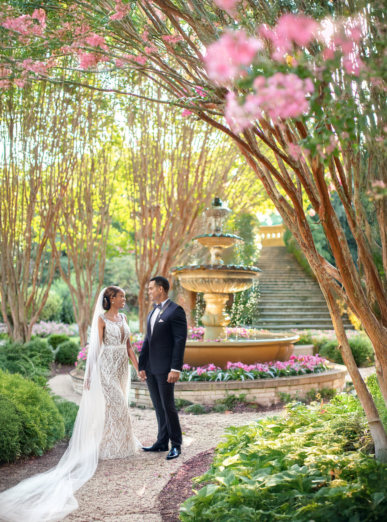 A bride and groom stand together in a lush garden, surrounded by blooming trees and greenery. A large, ornate fountain is in the background, with water cascading down its tiers. The bride wears a lace gown, and the groom is in a dark suit.