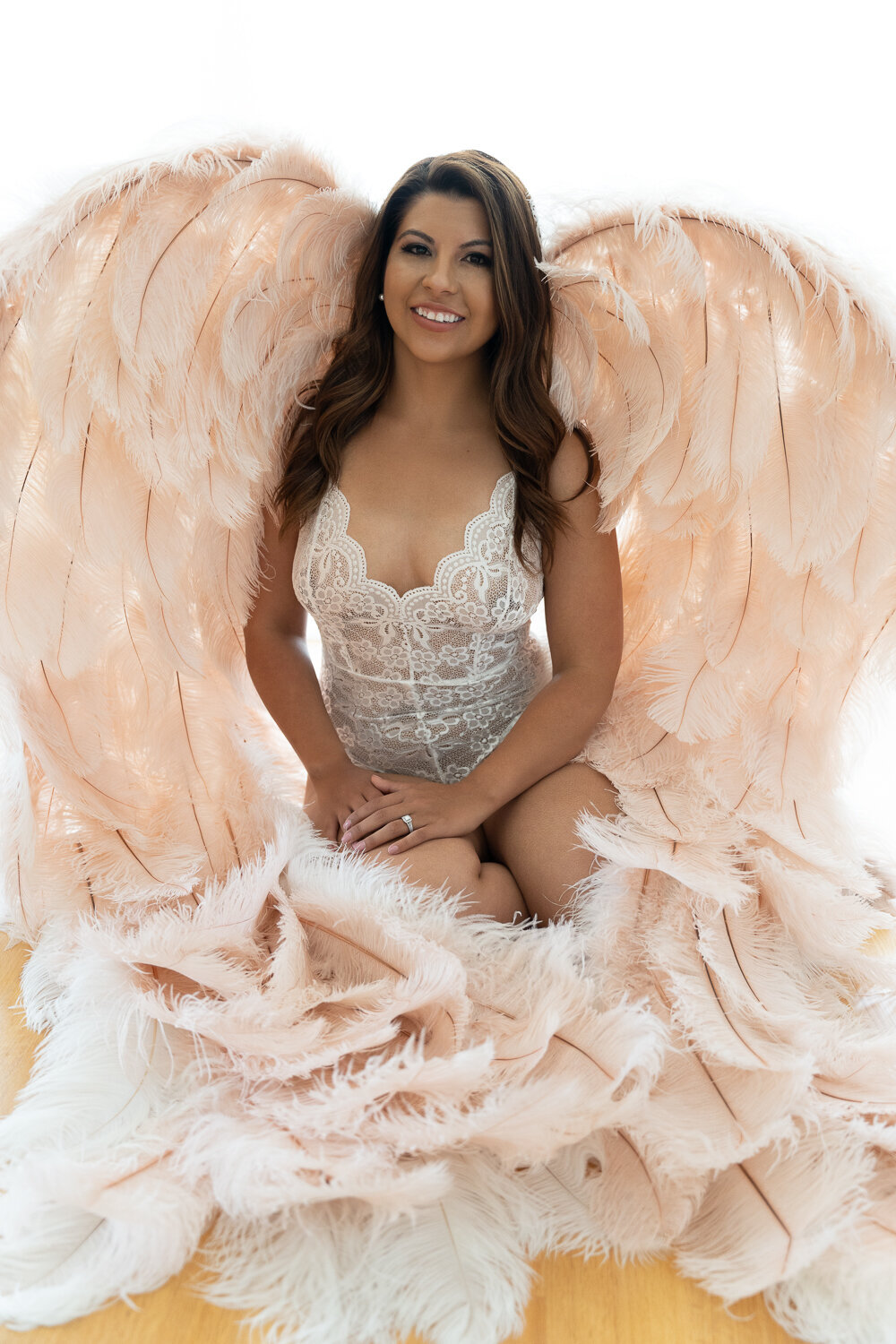 A radiant woman sits confidently on a bed in a Bay Area boudoir studio, adorned with large, plush feathered wings in a soft peach hue, complementing her lace bodysuit. Her joyful smile and the elegant wings create a celestial atmosphere