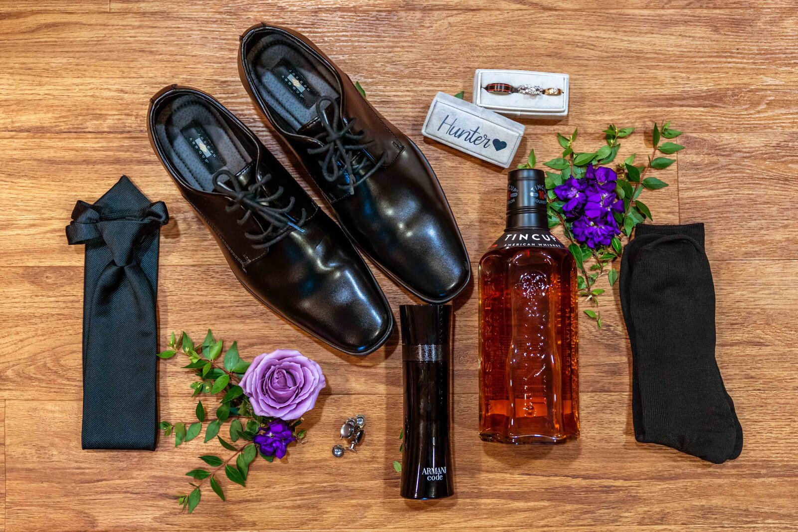A groom’s flat lay featuring polished black shoes, black bow tie, bourbon, cufflinks, and a boutonniere with purple accents, captured on wooden flooring during a Boone, NC wedding, showcasing elegant and personalized details.