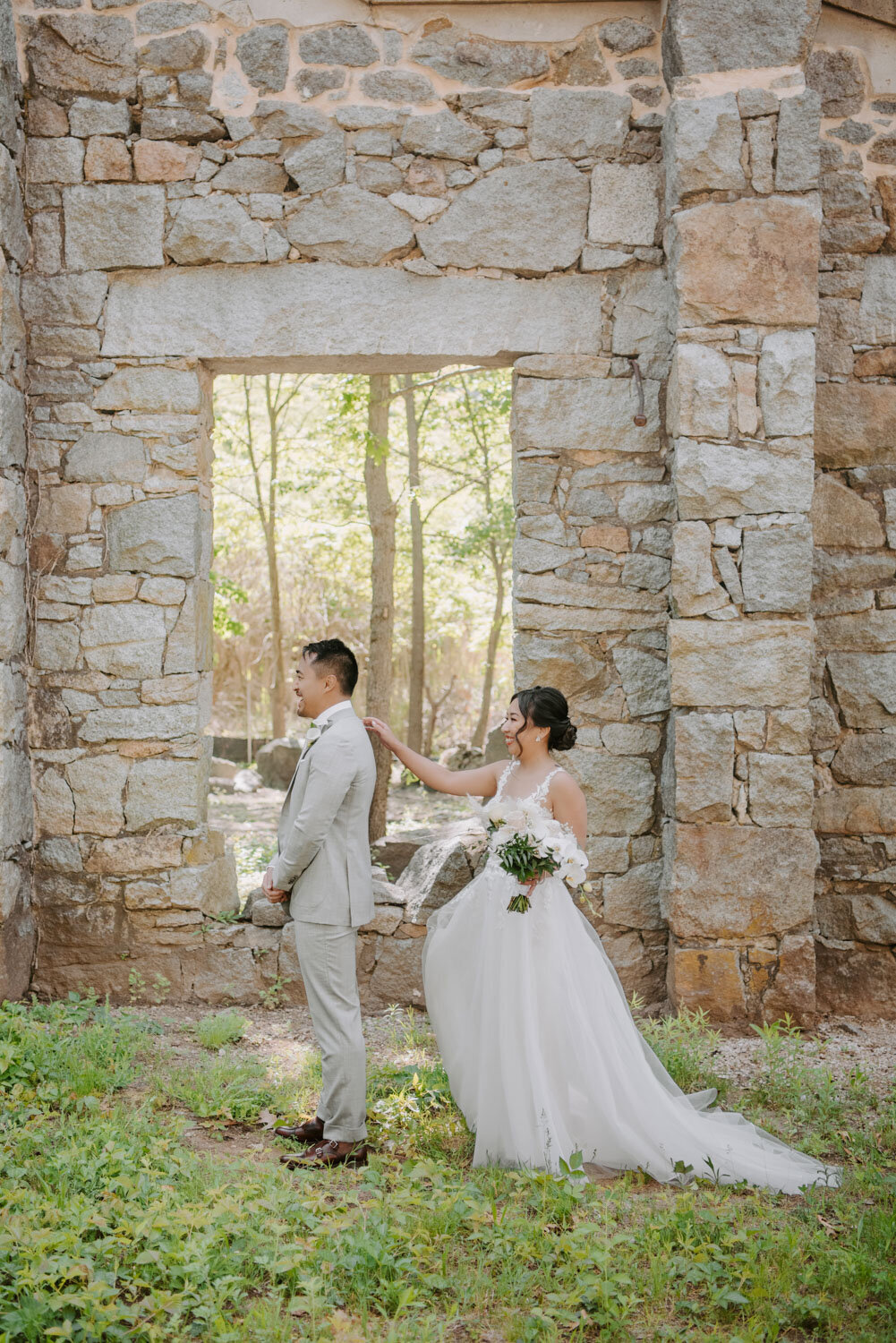 Bride tapping groom’s shoulder for a first look, surrounded by stone walls