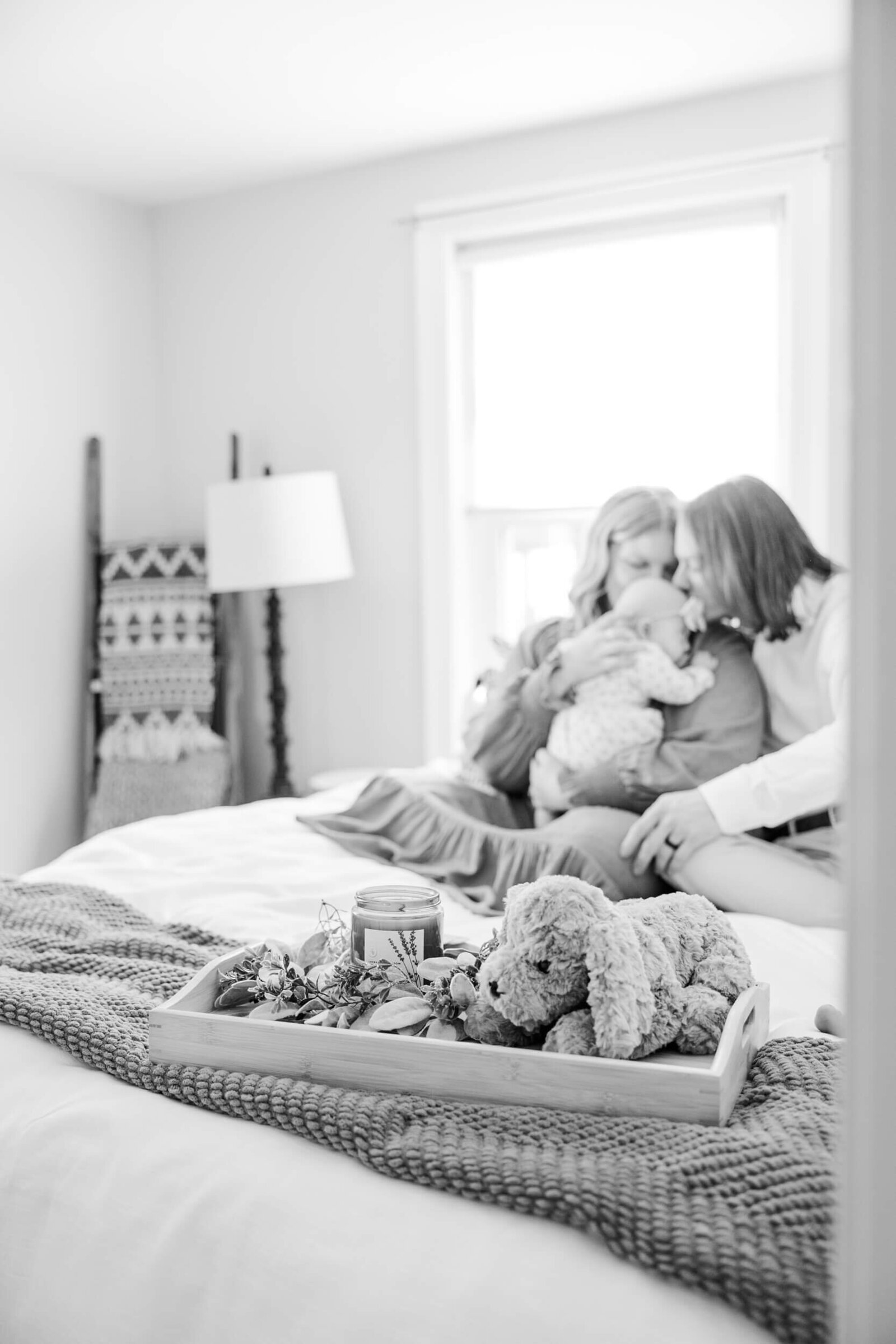 Black and white, focus is on a tray with a stuffed dog and candle; mom and dad kissing a newborn is soft and blurry in the background