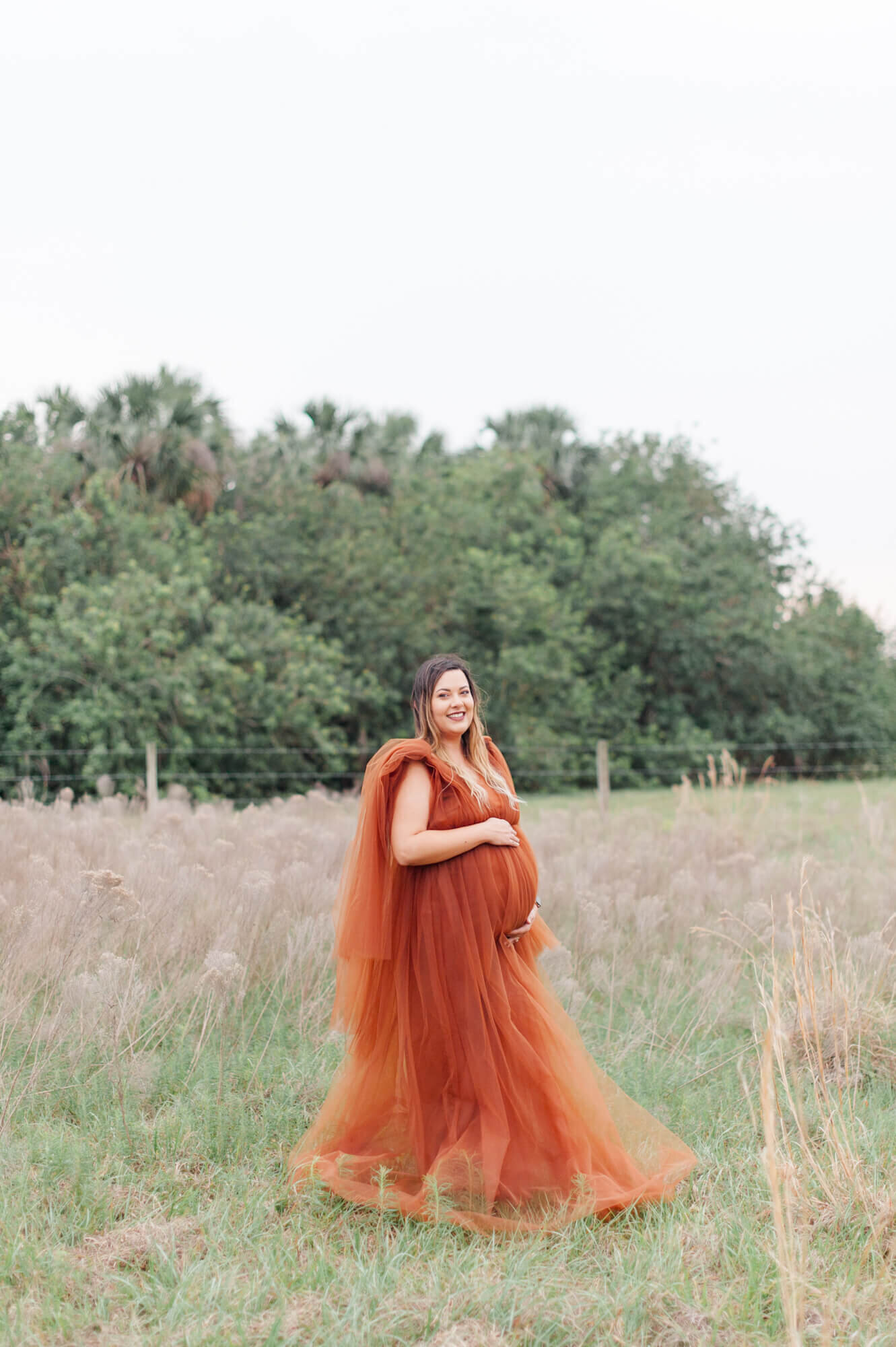 Expectant mother stands in a tall grass field wearing a beautiful tulle gown