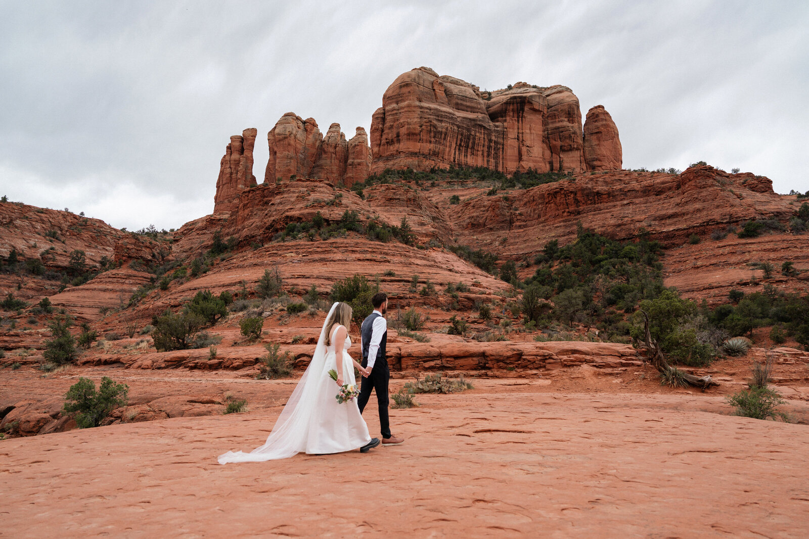 Bride and groom holding hands while walking across the red rock landscape during their elopement at Cathedral Rock in Sedona, Arizona, surrounded by dramatic cliffs and desert scenery.