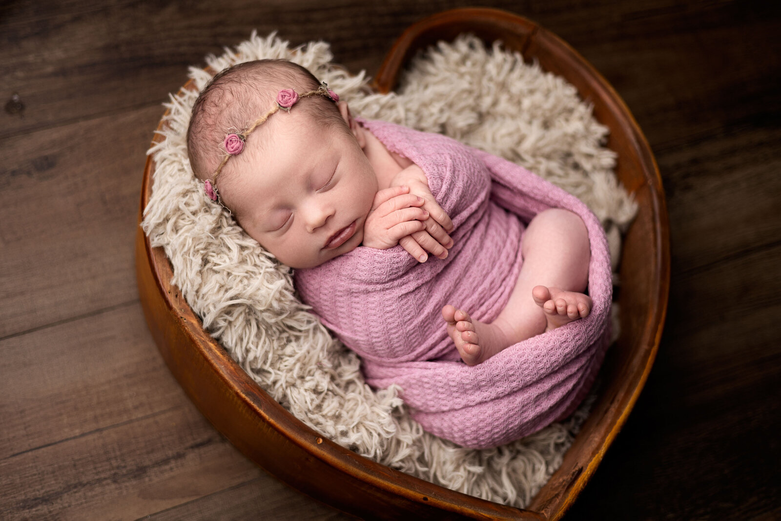 Baby girl wrapped in pink laying in heart shaped bowl with rose headband