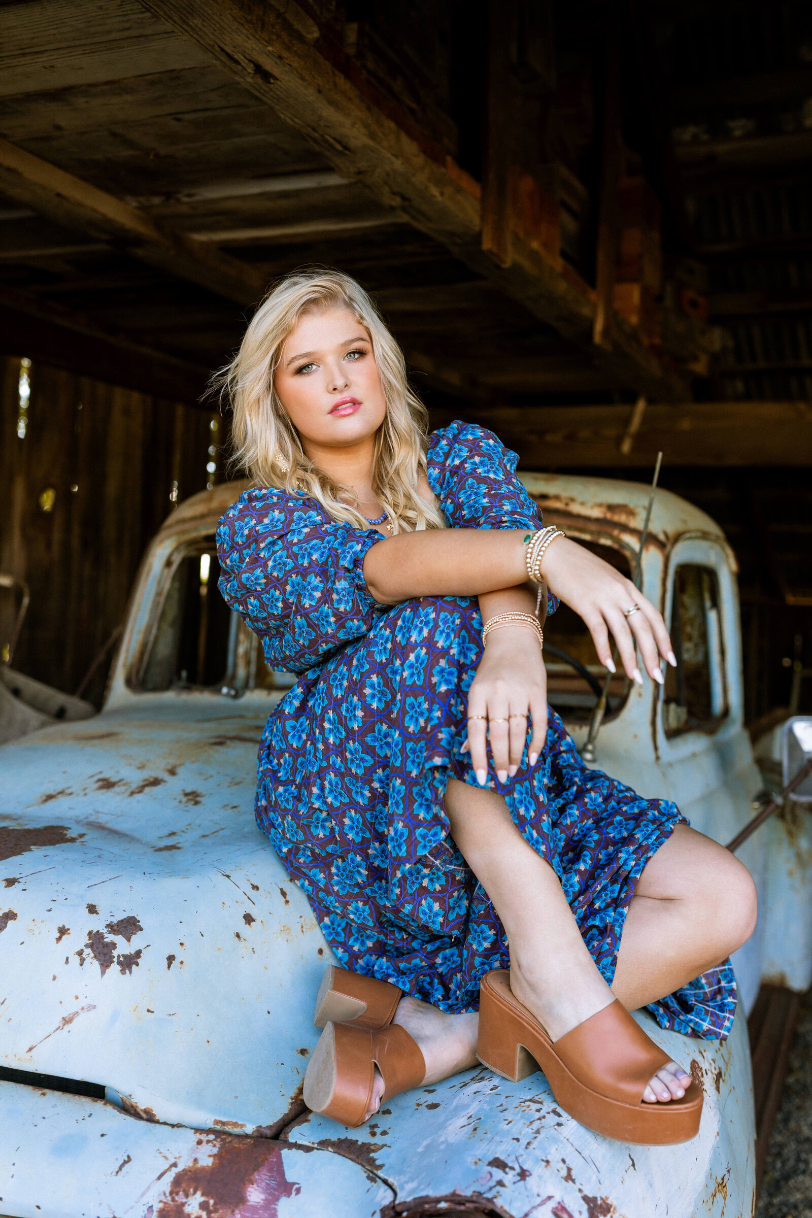 blonde girl wearing blue dress and brown heels sitting on top of an old rusted truck in a barn