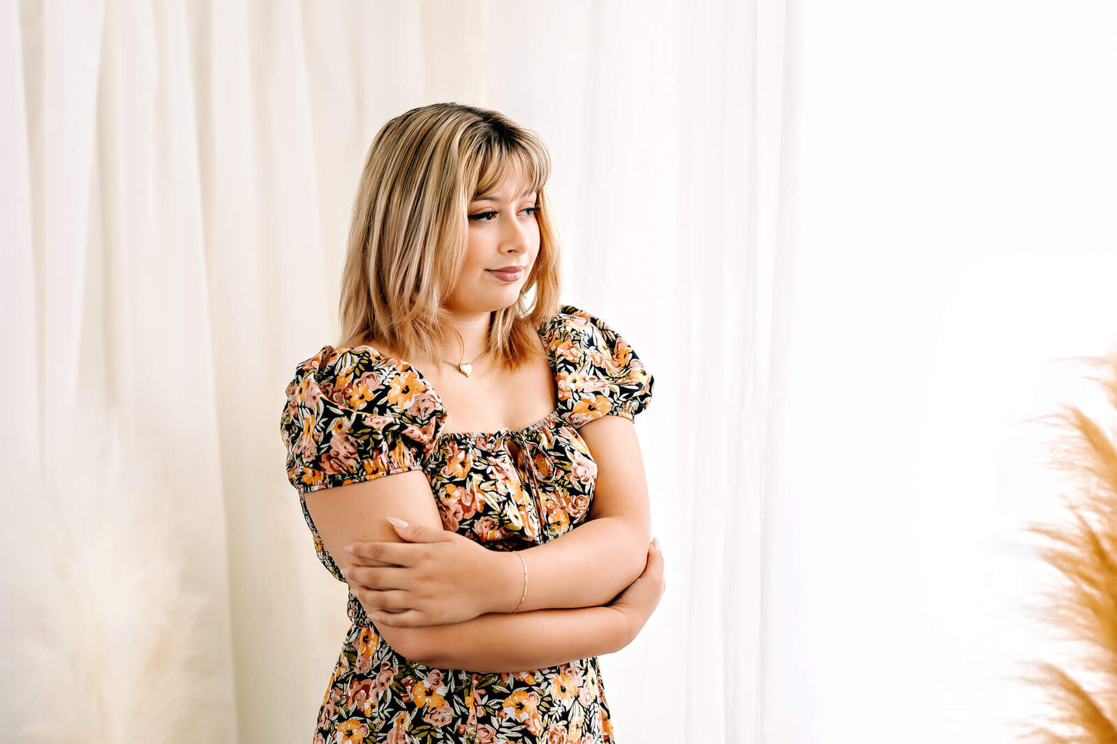 A person with shoulder-length hair stands with arms crossed, wearing a floral dress. The background is softly lit with sheer curtains and dried plants visible. They are gazing thoughtfully to the side.