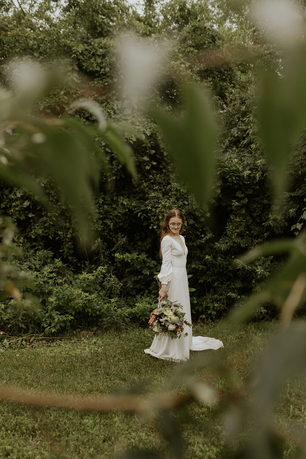 Bride standing alone in front of large green hedge while looking down at her green and white bouquet