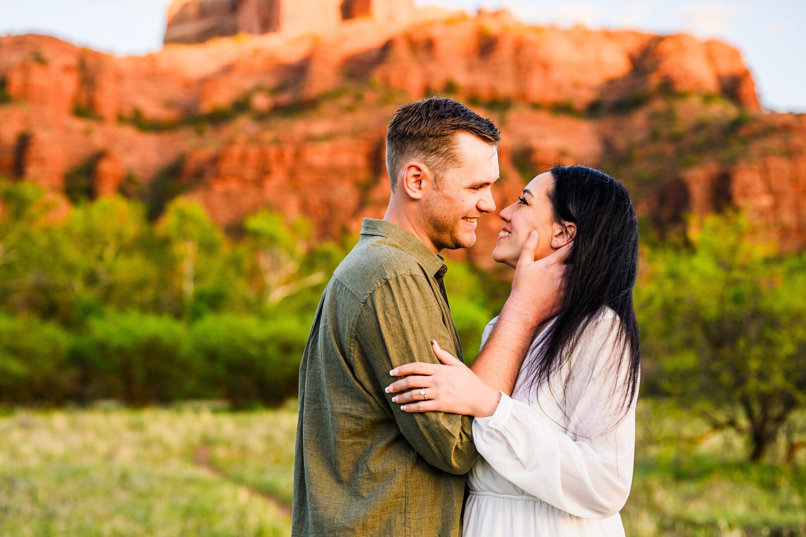 engaged in love couple looking at each other smiling holding each other with hands with red rock and trees background in Sedona at golden hour