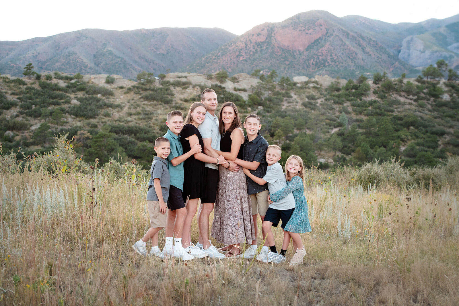 A family of 8 gather round mom and dad for hugs while standing in a field at sunset