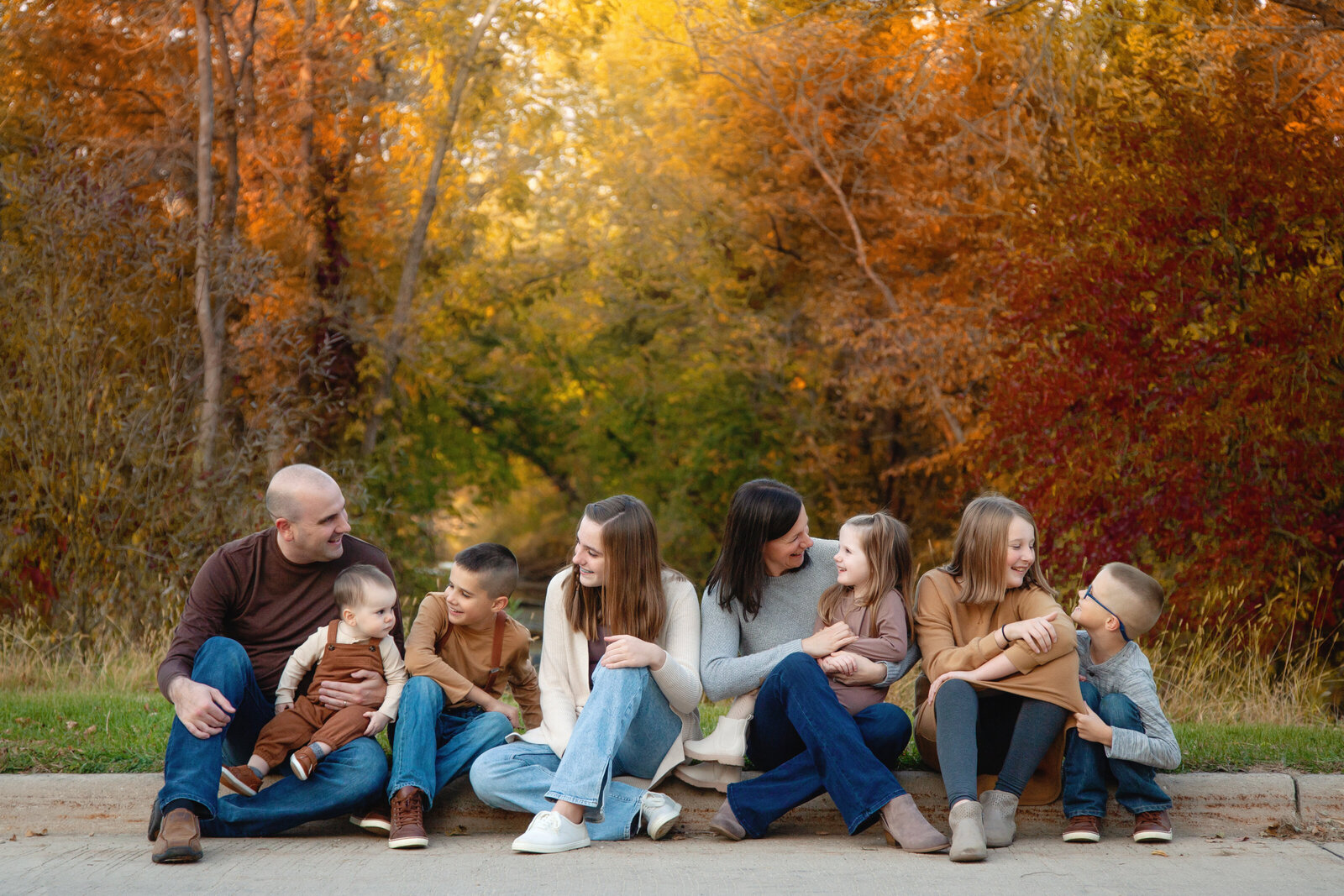 Family of eight sitting on curb smiling at each other, bright fall colors in background.