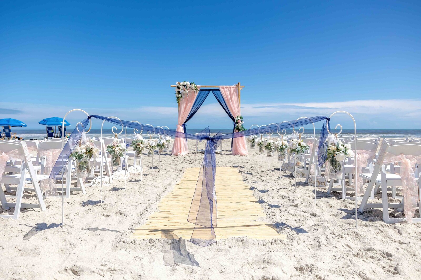 A beach wedding ceremony setup with a pink and navy draped arbor, white chairs, and floral accents, captured during a Topsail Beach, NC wedding, blending coastal elegance and vibrant details.