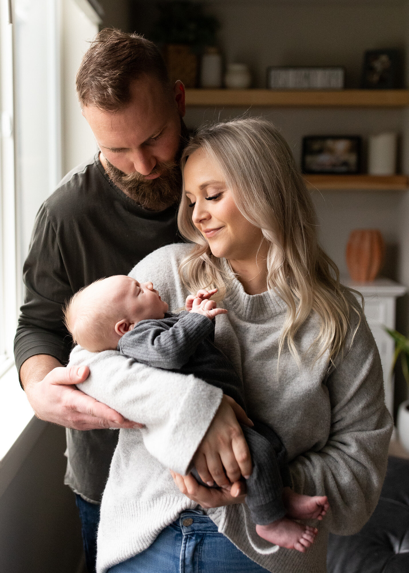 Parents of newborn holding him by a naturally lit window.