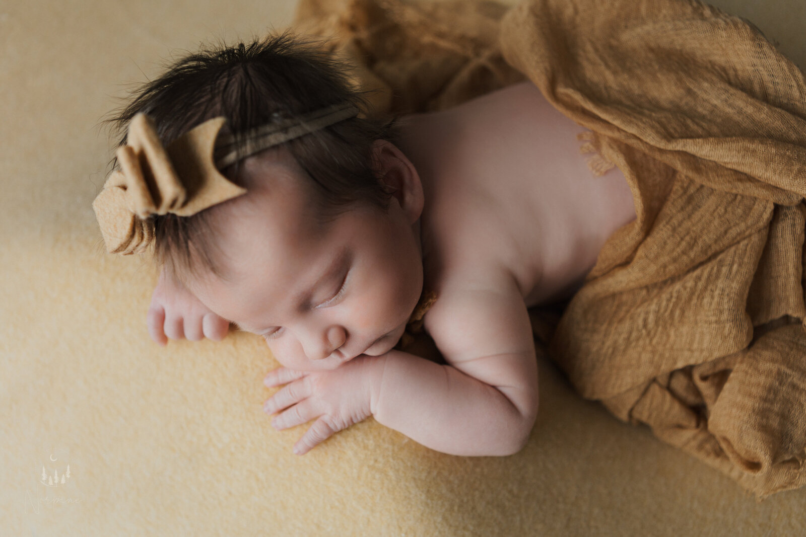 a newborn girl with a yellow wrap, backdrop and headband laying on her stomach with her hands near her face, sleeping. The image is taken from above to show her side profile and back.