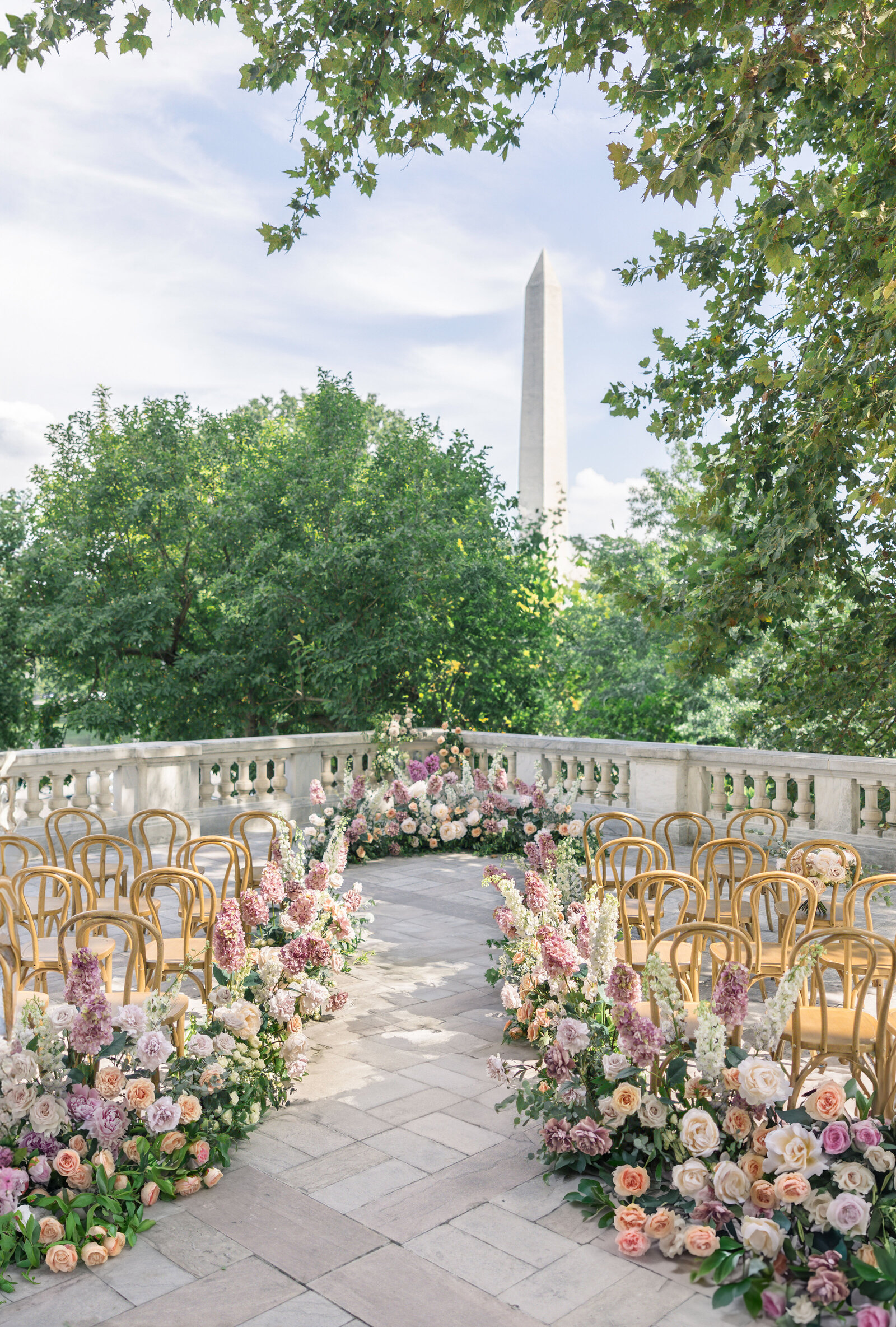 A beautiful outdoor wedding setup with rows of wooden chairs and lush floral arrangements. In the background, a tall monument rises above the greenery, under a partly cloudy blue sky.