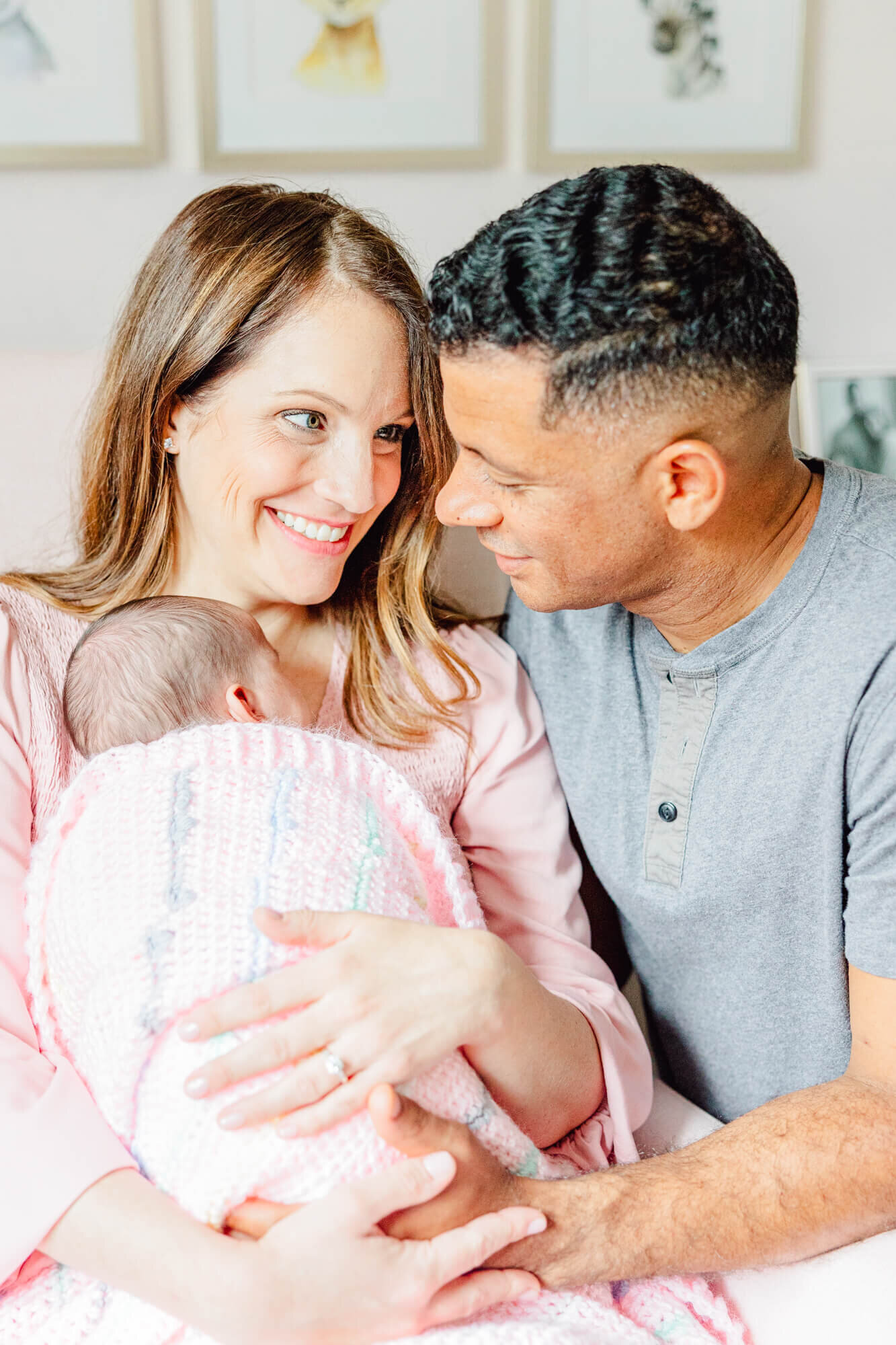 Mom and dad smile together and hold their newborn in a knitted pink blanket