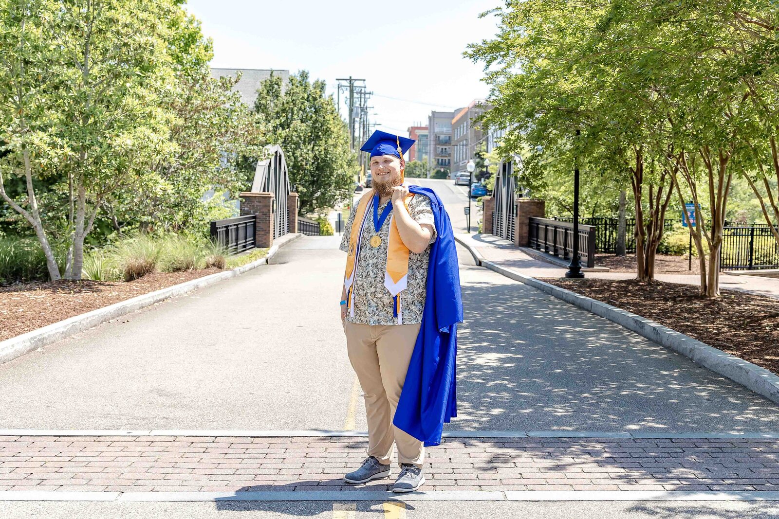 A CFCC senior in a blue cap and gown standing proudly on a bridge, holding the gown over his shoulder in Wilmington, North Carolina. This photo highlights graduation excitement, ideal for capturing vibrant senior photography in an urban setting.