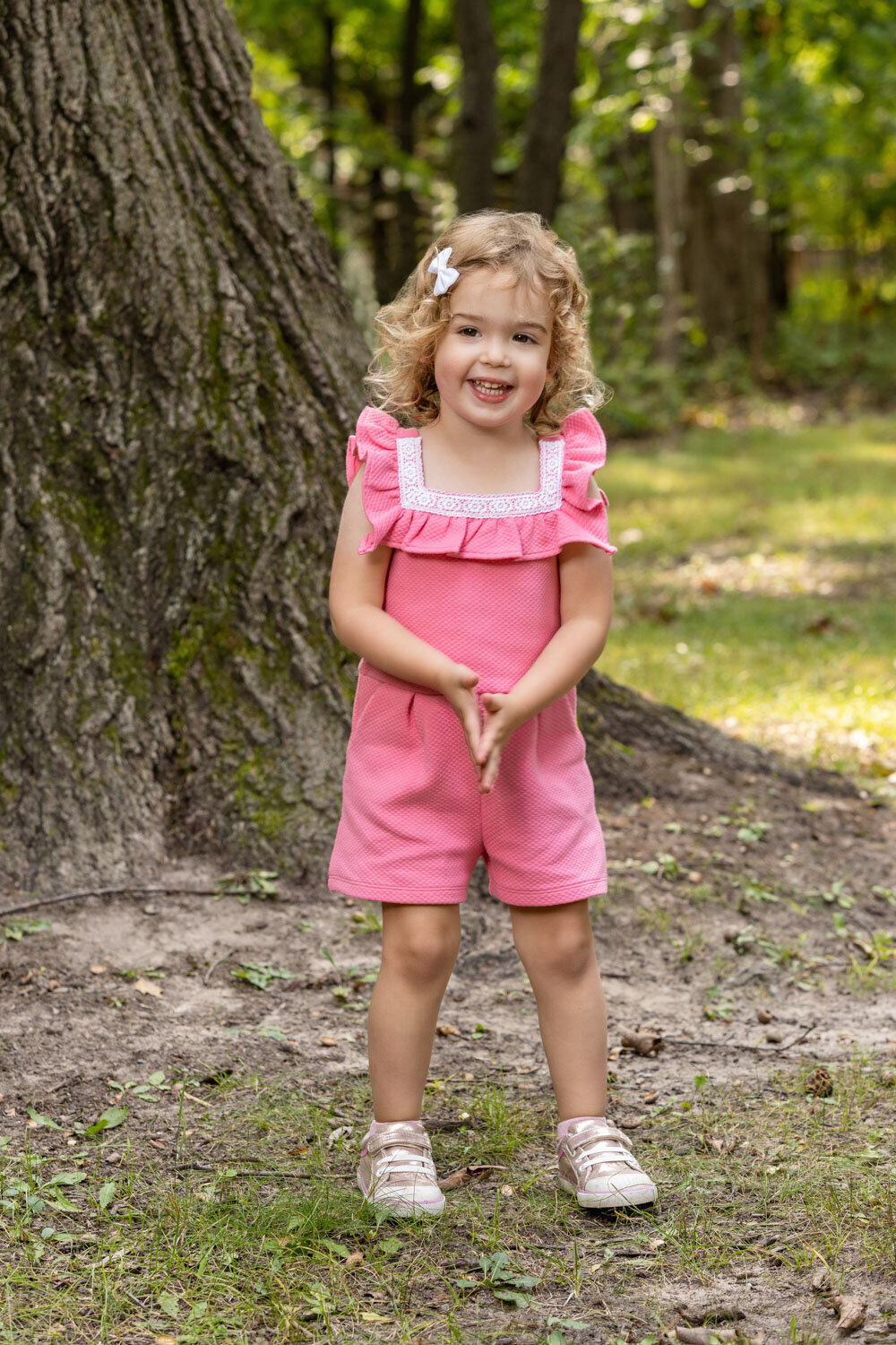 A little girl in a pink jumpsuit is standing in front of a tree.