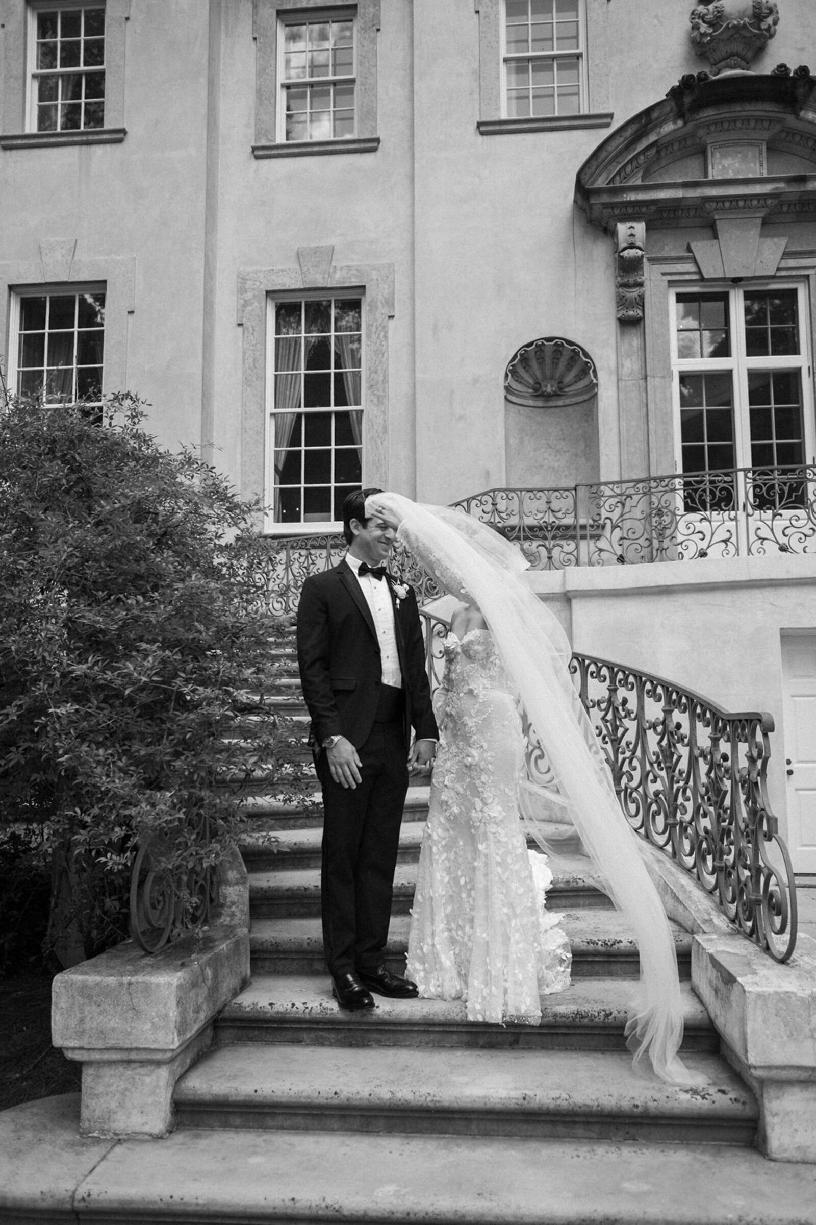 Candid black & white portrait of bride & groom standing on outdoor steps of European-style mansion with bride swooshing veil in front of their faces 