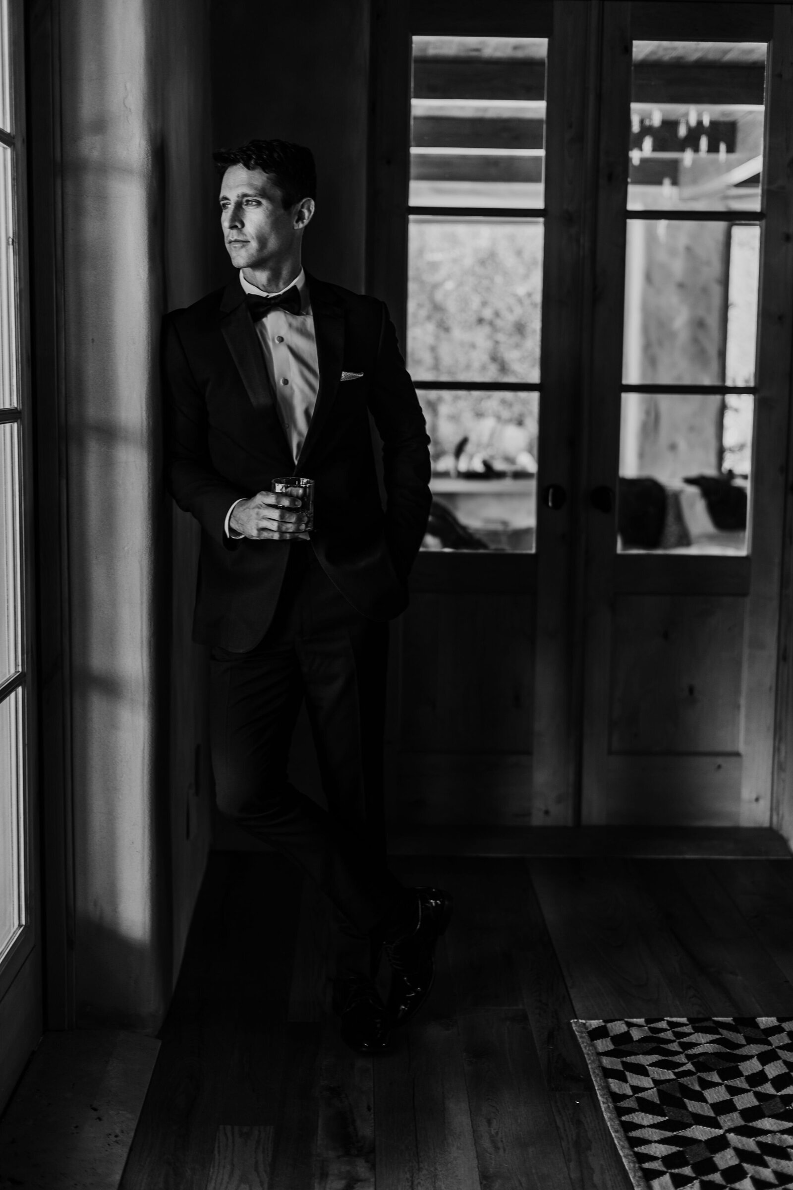 black and white wedding portrait of groom leaning against a wall as looking out a floor to ceiling window while holding a drink in his hands