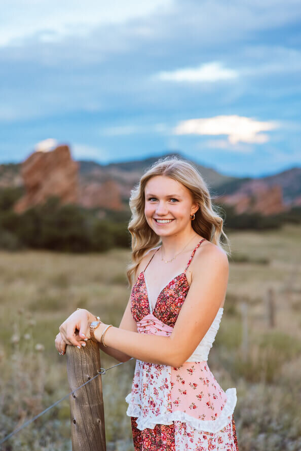 Girl in pink flower dress poses for senior photos with red rocks behind her.