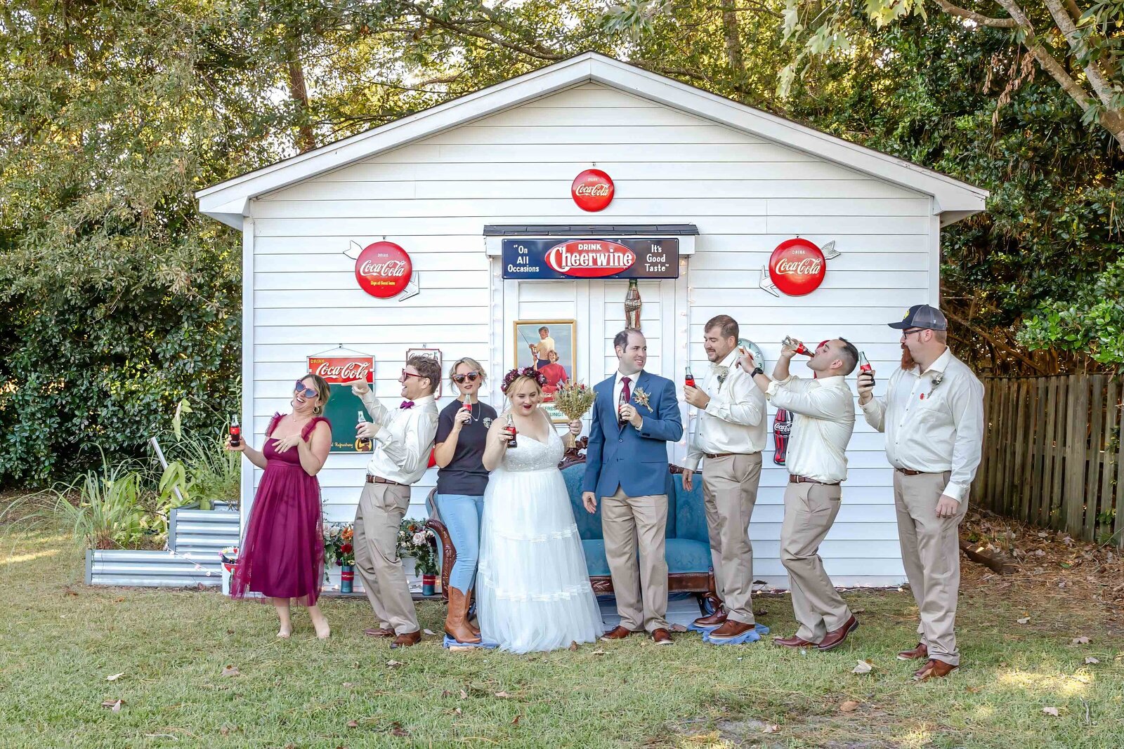 The bride, groom, and wedding party pose playfully in front of a Coca-Cola-themed vintage shed with colorful decor, captured during a Wilmington, NC wedding, showcasing unique and fun wedding details.