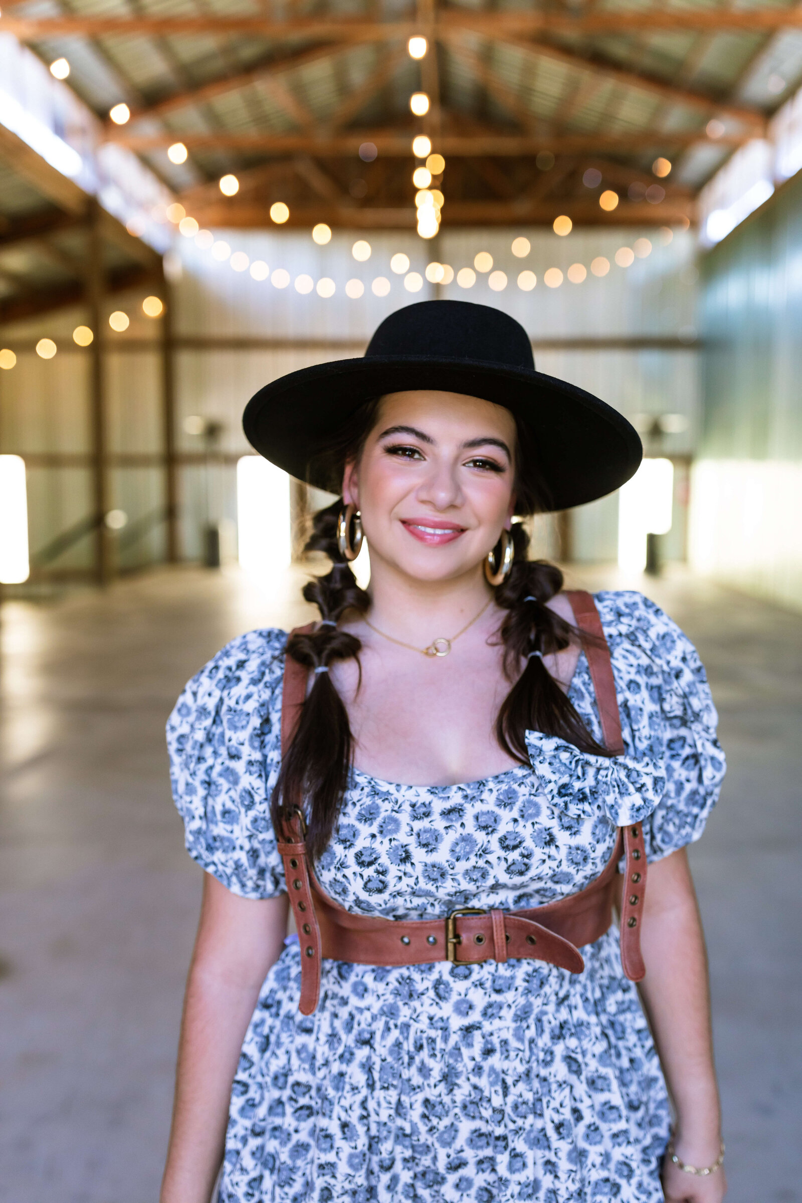 dark haired girl wearing black hat, brown belt, and blue floral dress with bubble braids in a warehouse