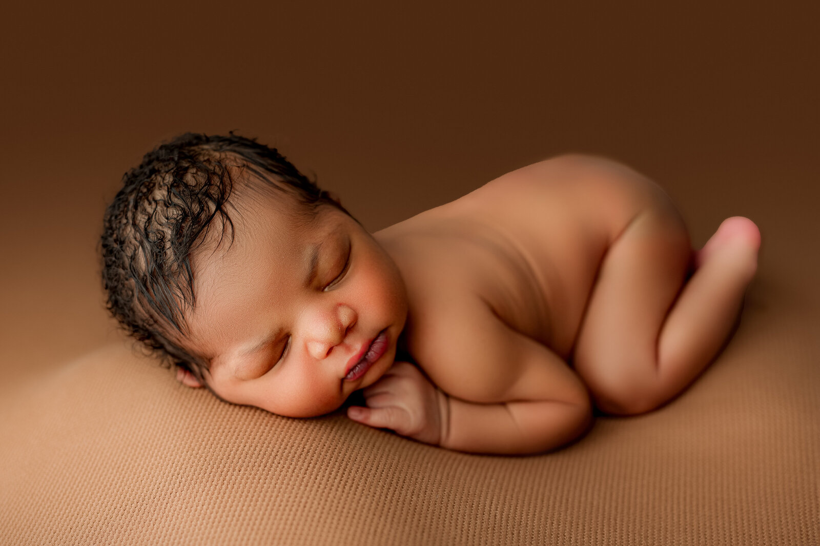 A newborn baby peacefully sleeps on a soft, brown surface. The baby is lying on their side with hands tucked under the chin, eyes closed, and a serene expression. Expertly captured by Waxhaw newborn photography, the warm brown background creates a cozy atmosphere.