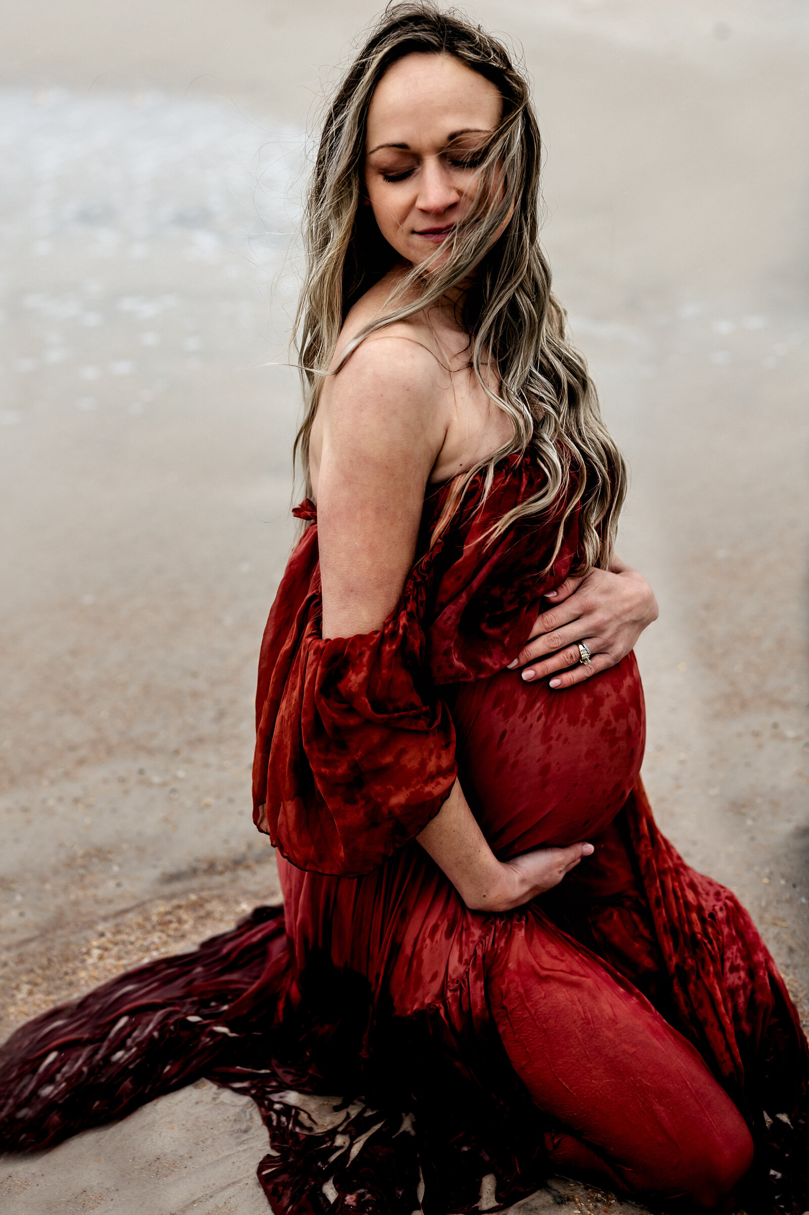 Pregnant woman in Golden Hour Gowns red dress kneeling in the ocean in Florida