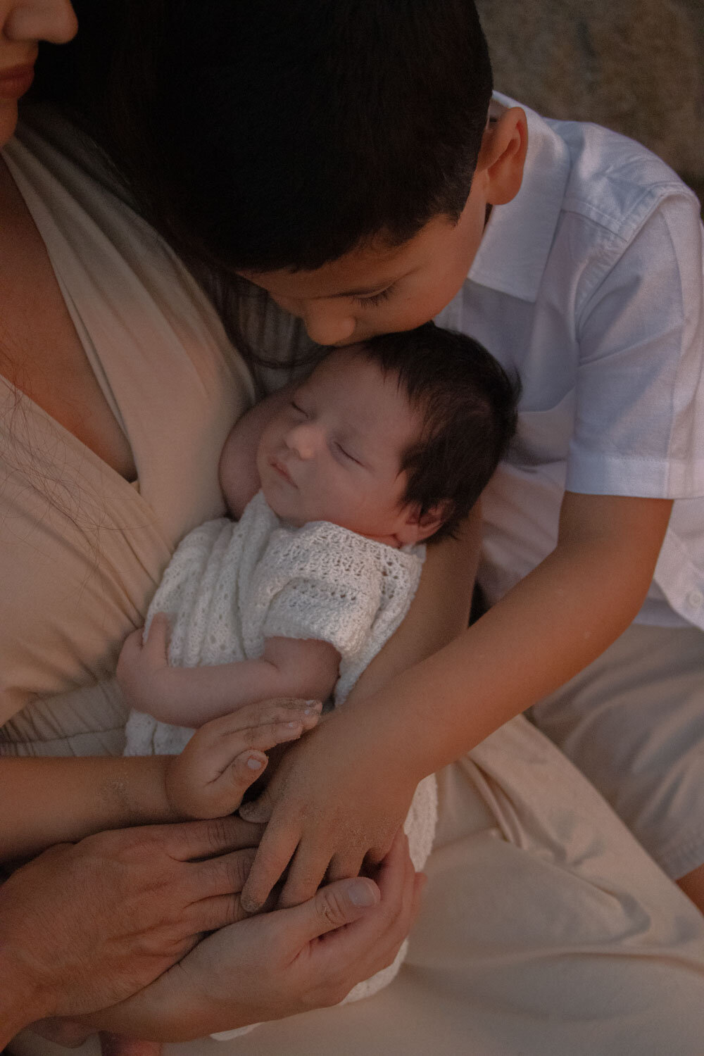 brother gently kisses his sleeping newborn sibling, held by their mother