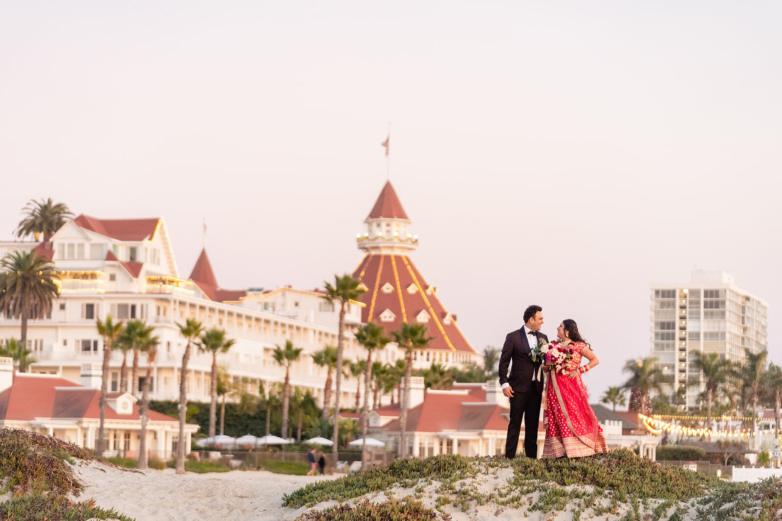 A groom in a black suit and a bride in a red and gold sari with the Iconic Hotel Del in the background