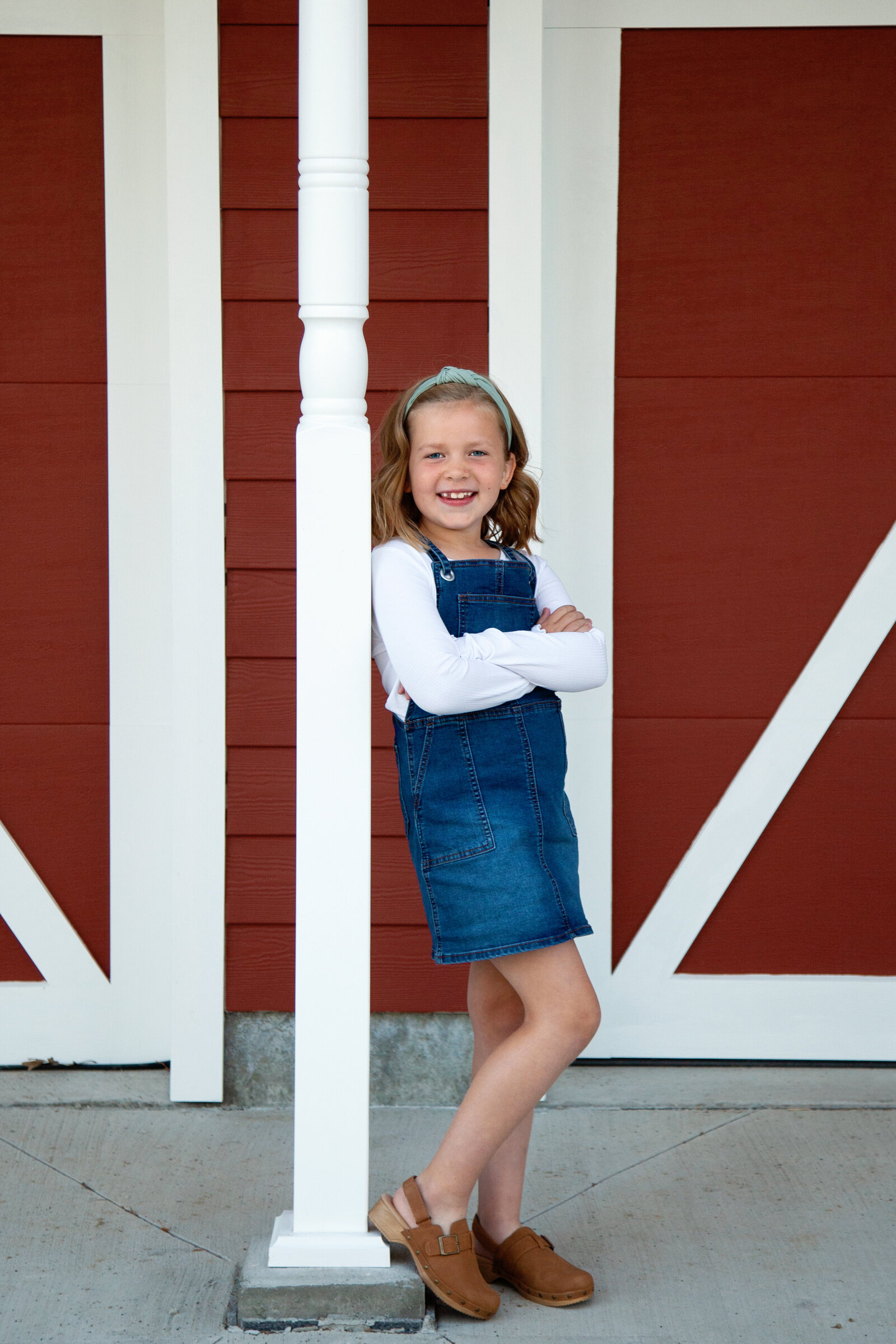 Girl in denim pinafore, arms crossed, smiling, red barn background.