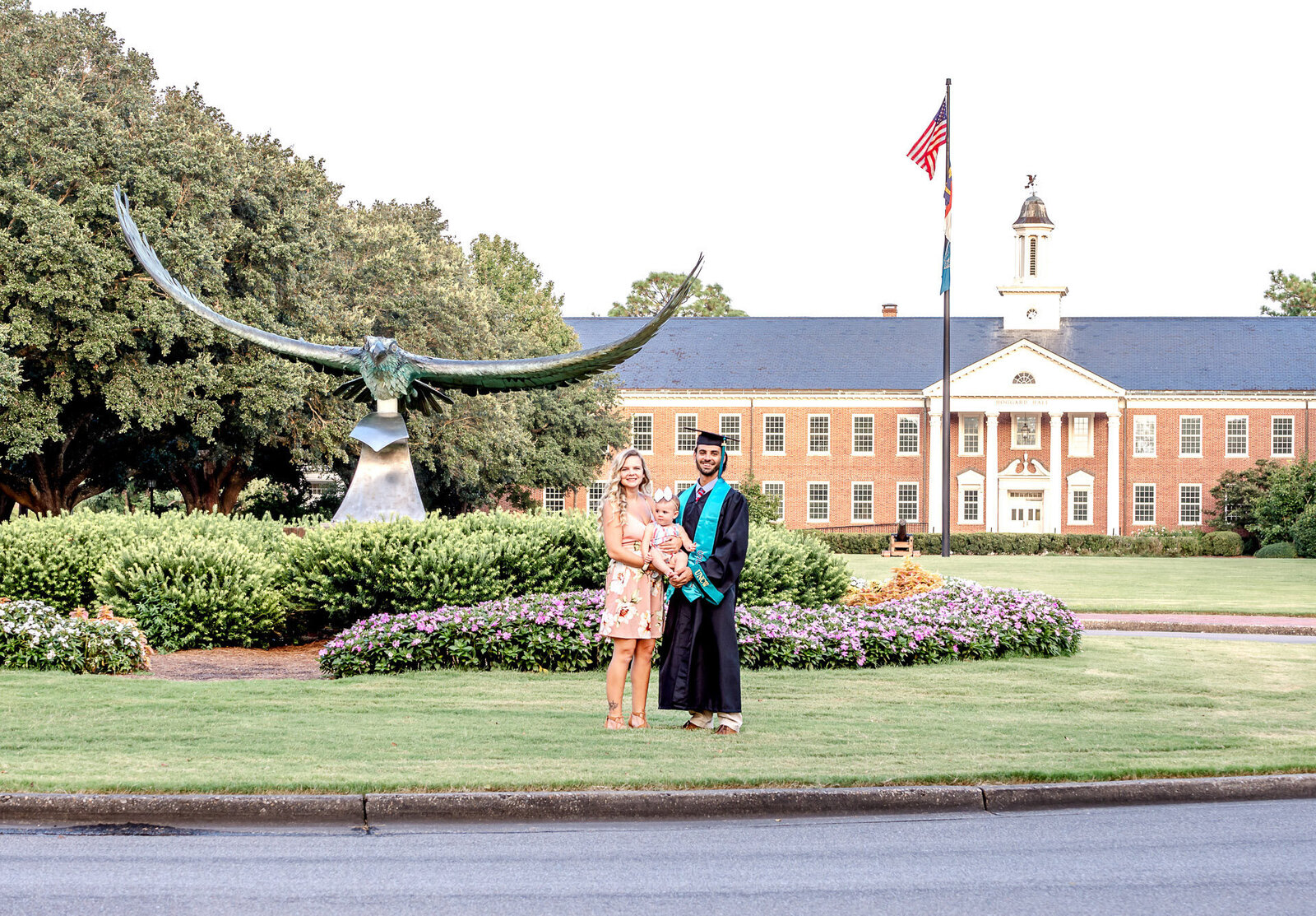 A UNCW college graduate in academic regalia standing with family on a campus lawn in front of a university building in Wilmingotn, North Carolina. This image captures the joy of graduation, perfect for capturing memorable family moments.