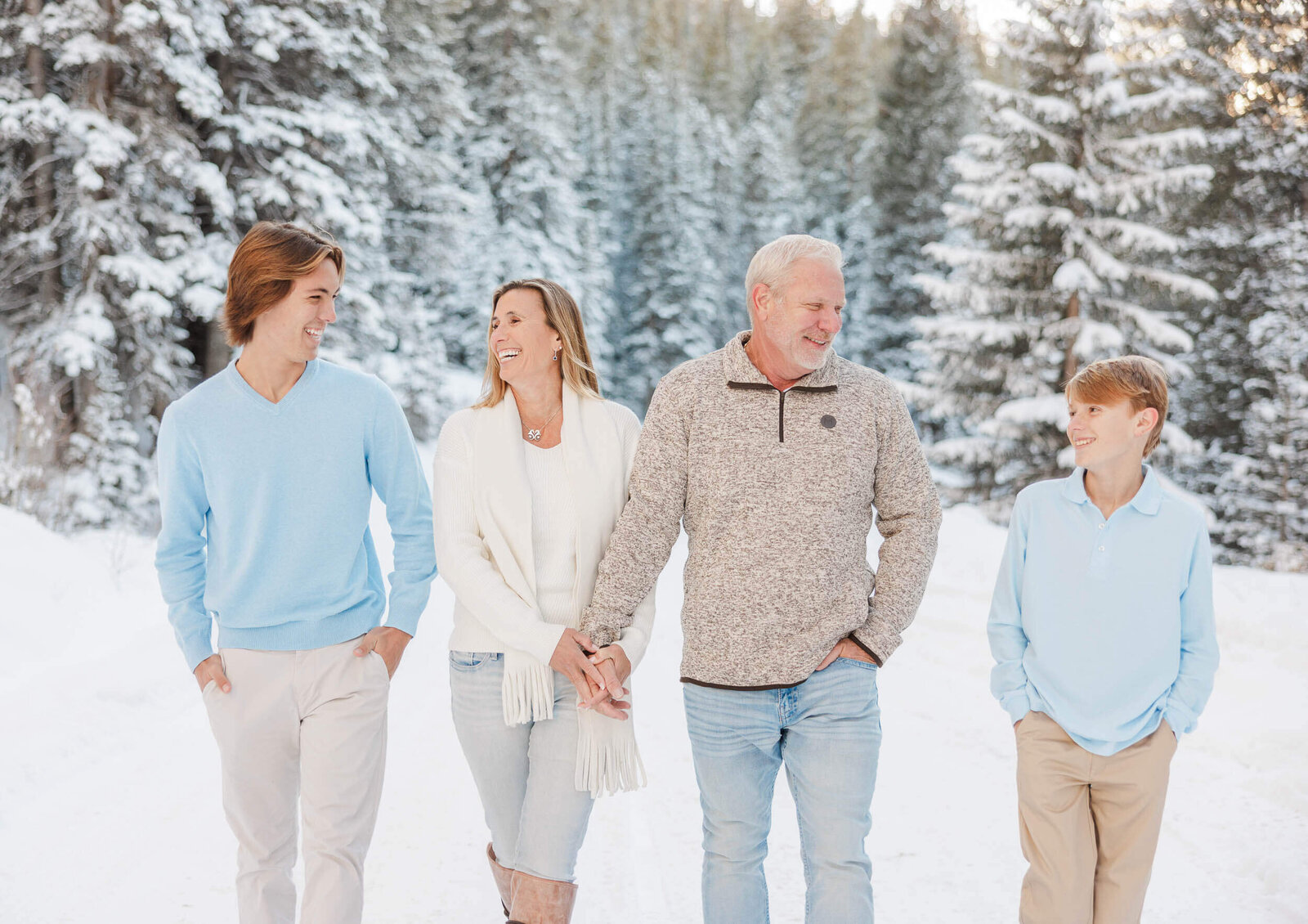 A family of four is walking hand in hand amongst a snowy winter scene. Surrounded by snow covered evergreen trees