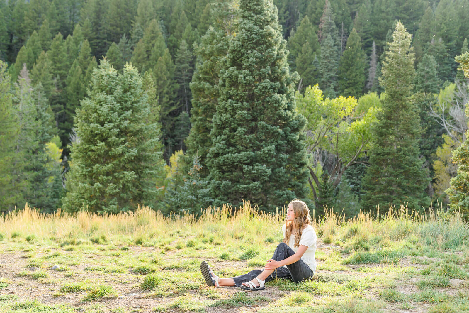 Salt Lake City photography of a high school senior girl wearing a white short sleeved blouse and dark jeans sitting in a grassy field with pine trees in the distance. Photo captured at Tibble Fork Reservoir in the Spring