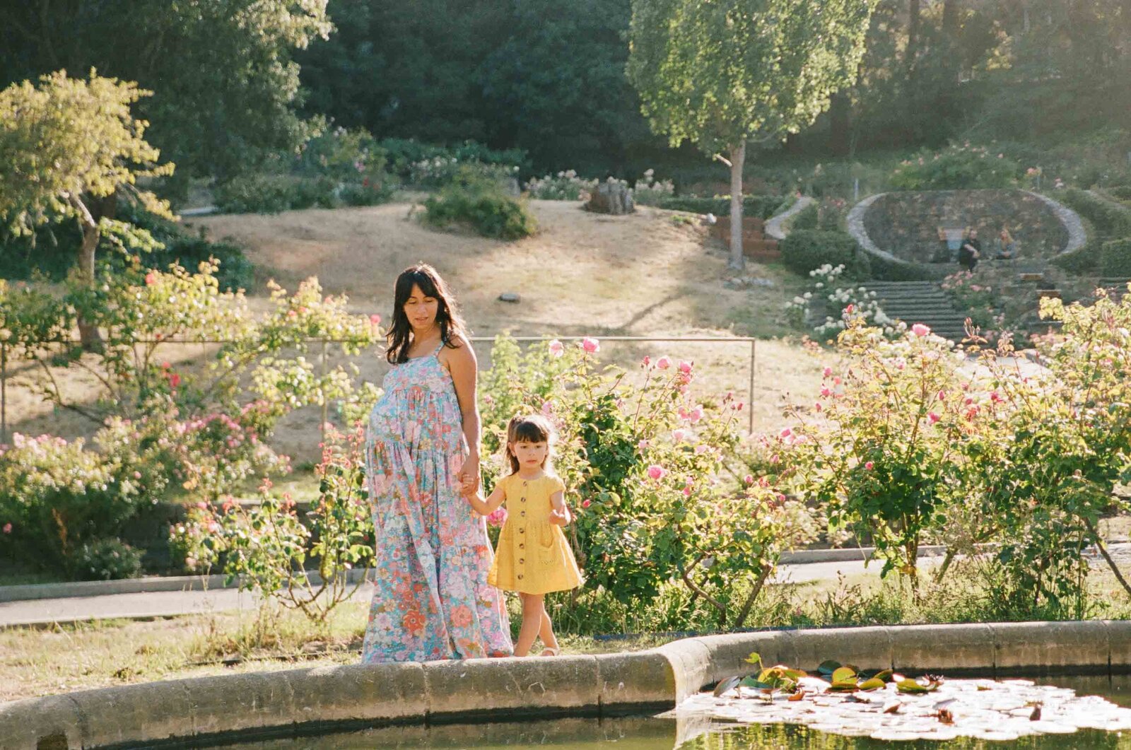 film photo of pregnant woman in a free people maxi dress holding hands with toddler daughter in yellow dress and walking in an oakland california rose garden