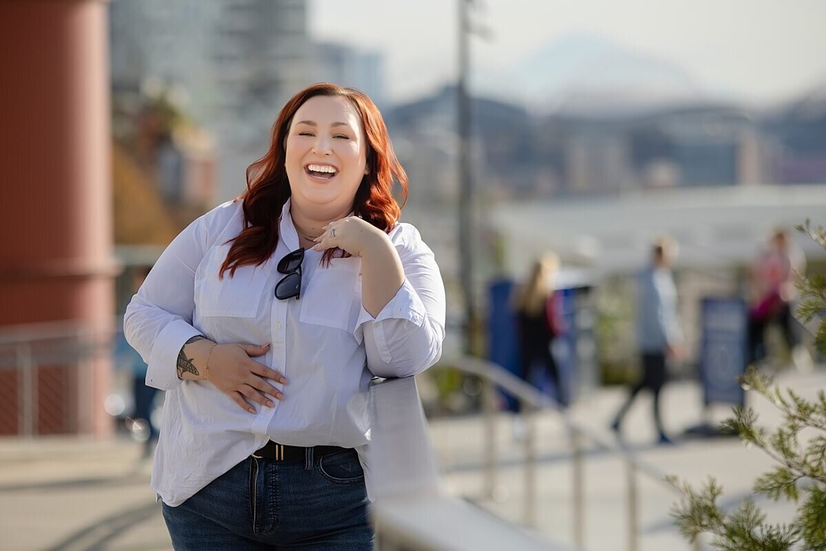 Personal branding photography of a joyful red-haired woman in a white shirt and jeans, laughing confidently during an outdoor Seattle photoshoot with cityscape and Mount Rainier in the background. Captured for “She Plays With Fire” by a Seattle brand photographer, showcasing authentic, approachable visuals for bold women in business.
