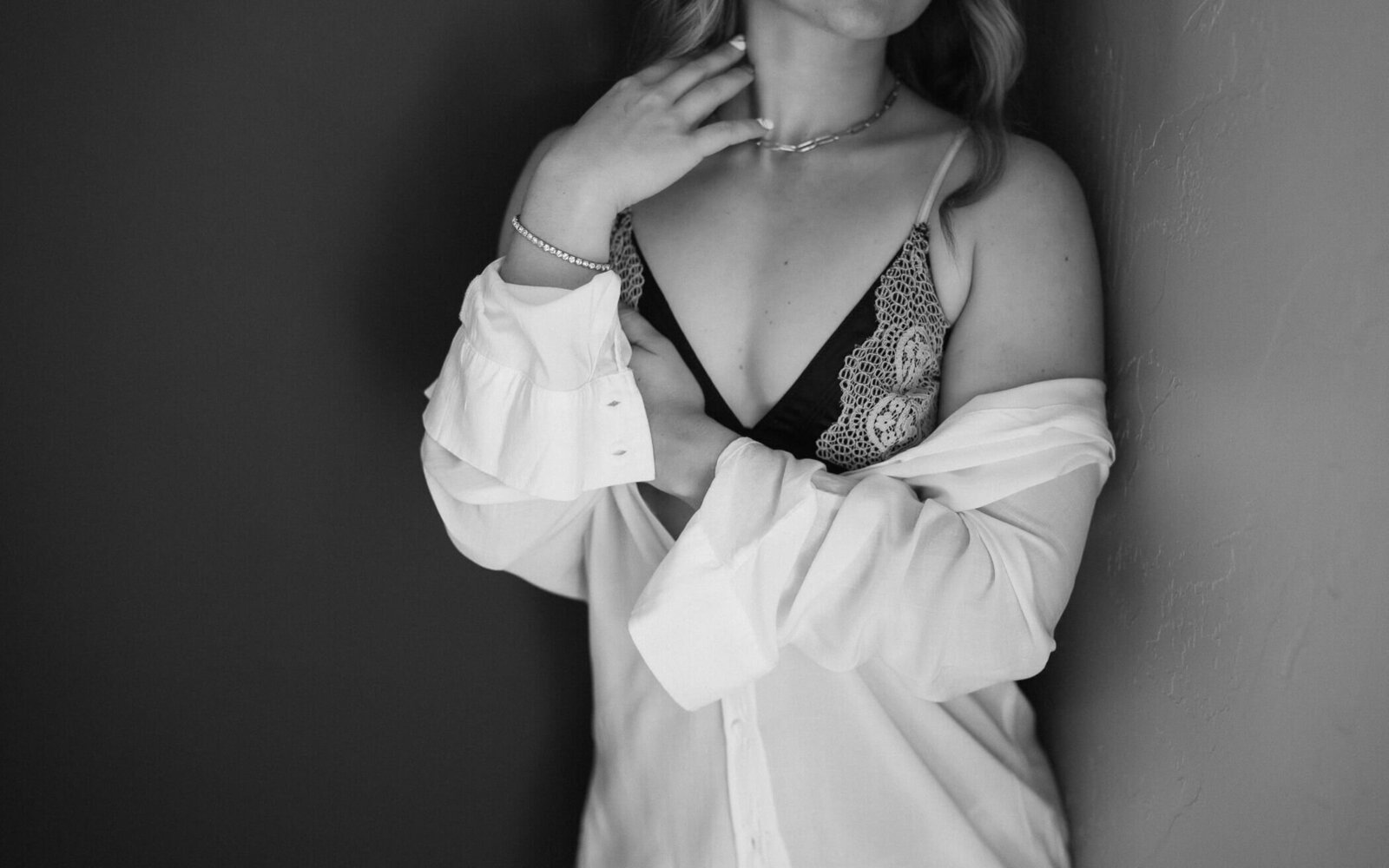 A close-up portrait that is indistinct of a woman wearing a lacy, black top, and an oversized unbuttoned white dress shirt, leaning up against a black wall.