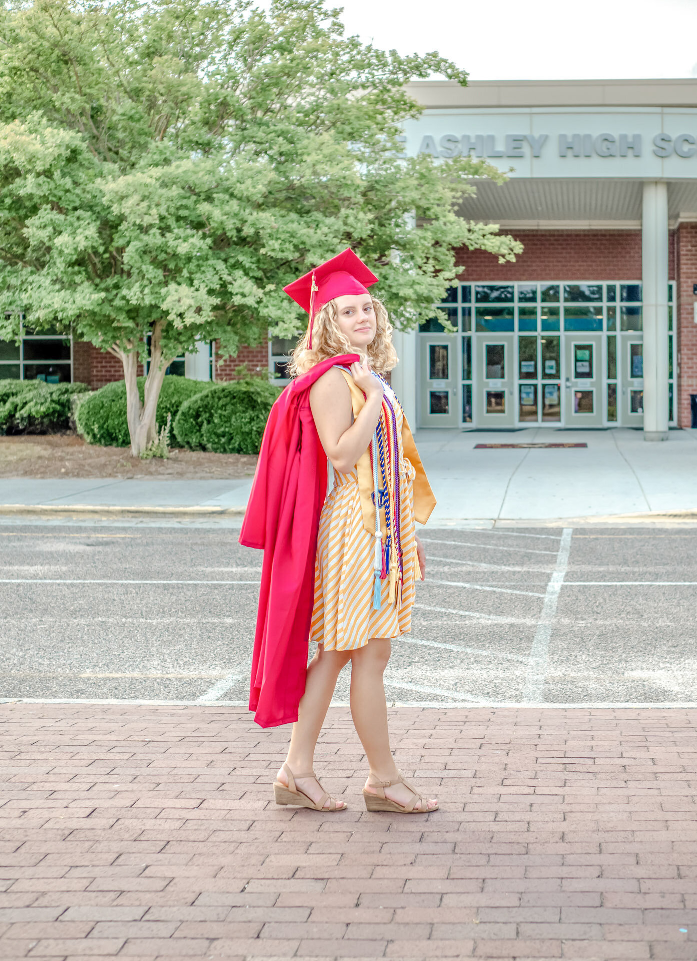A high school graduate in a red cap and gown holding a diploma in front of a large Ashley High School building in Wilmington, North Carolina. This image captures the pride of graduation, ideal for those seeking professional senior photography in an academic setting.