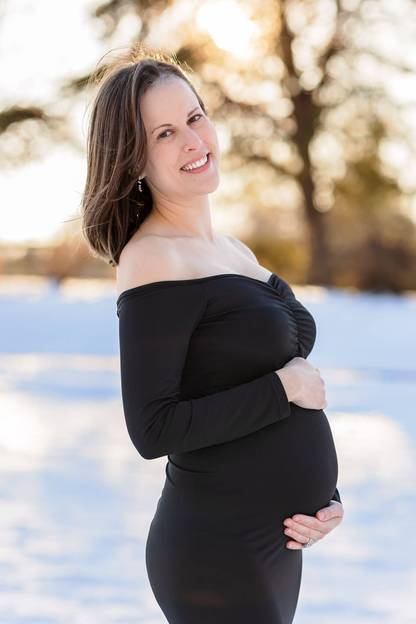 A pregnant woman in a black dress hugging her belly while posing in the snow at Burke Lake Park.