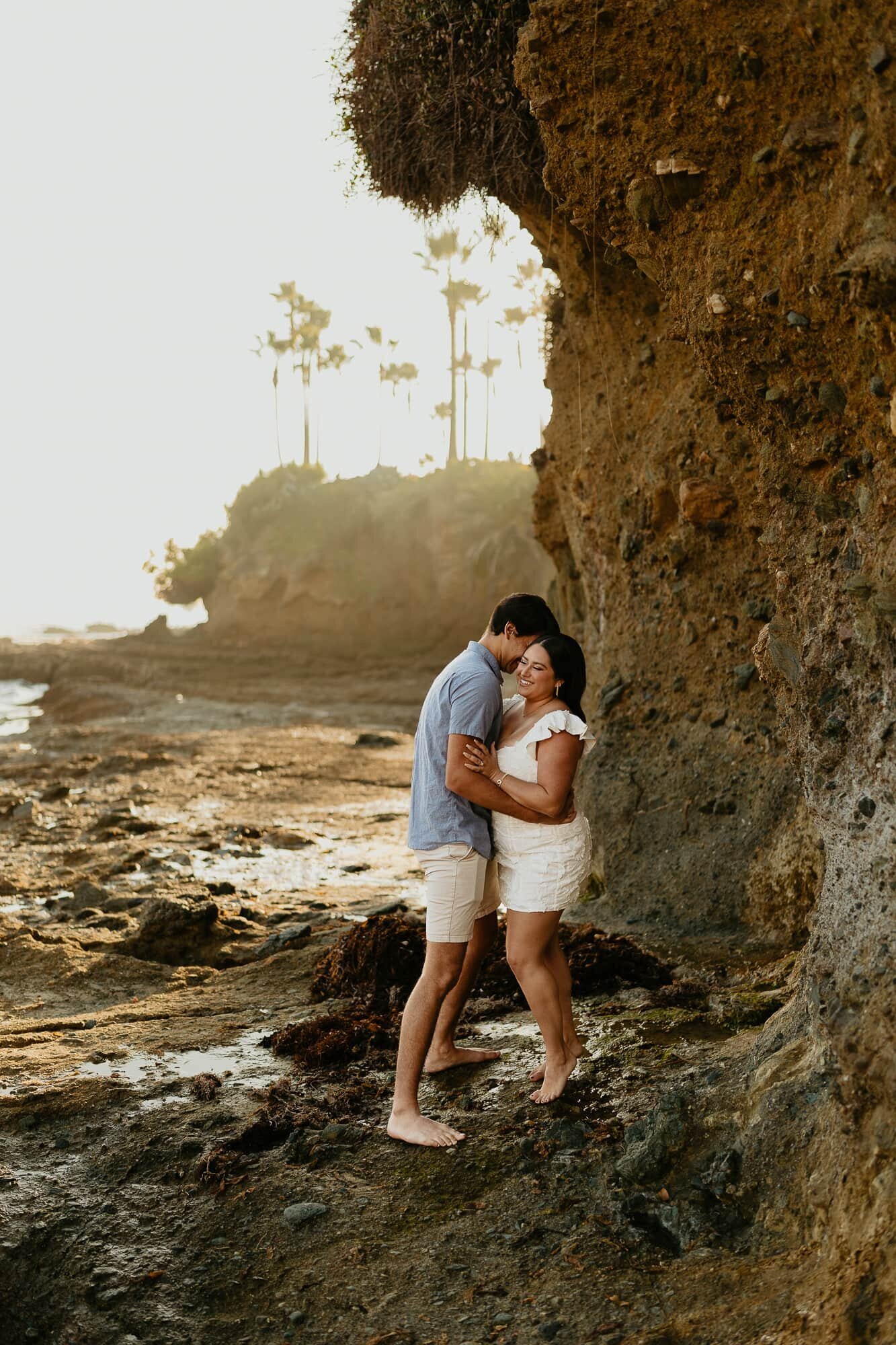 A couple standing under a cliff during their engagement photo session in Laguna Beach, California