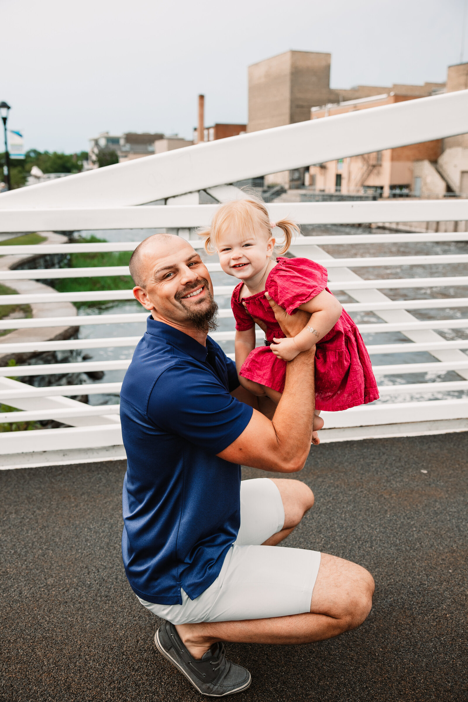 A happy father squats and lifts his toddler daughter in a pink dress on a modern bridge