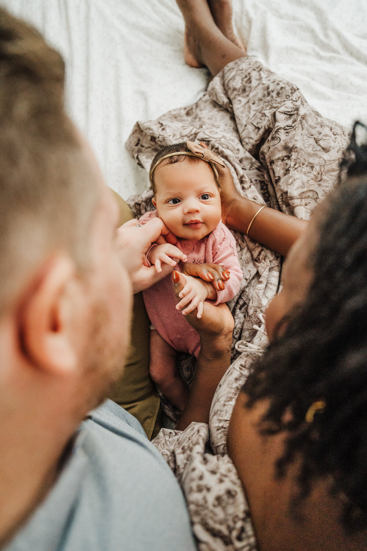 camera peeks between the heads of two parents to see newborn girl looking at camera