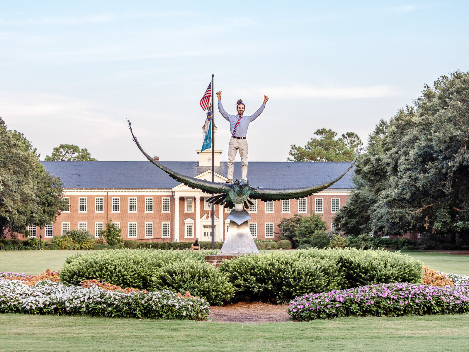 A graduating senior in a tie standing triumphantly on the seahawk statue in front of a University of North Carolina Wilmington building in Wilmingotn, North Carolina. This dynamic photo celebrates academic achievement, perfect for capturing memorable senior moments on campus.