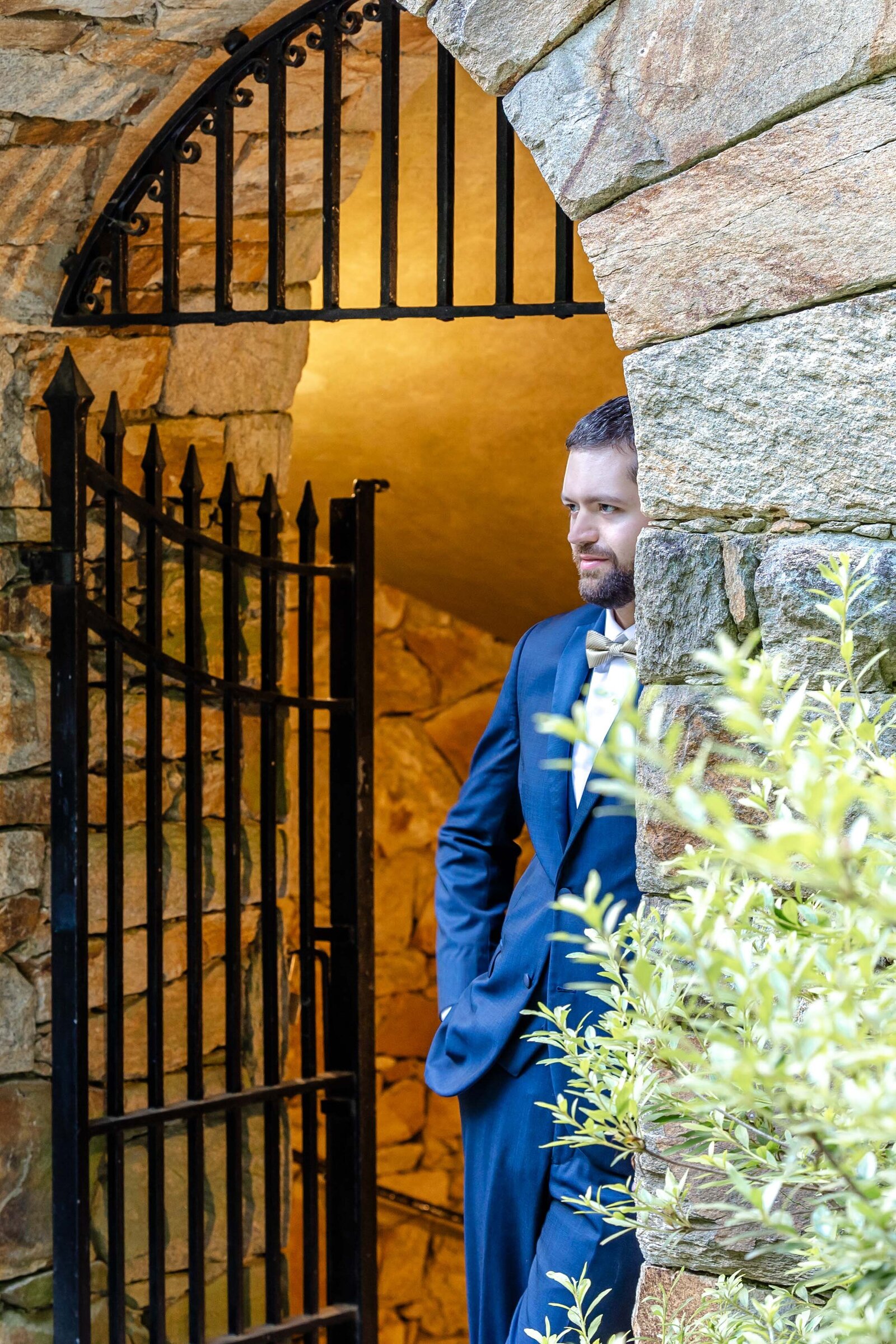 A groom in a navy suit poses by an arched stone gate with iron detailing, surrounded by greenery, captured during a rustic and elegant Longwood Gardens, Kennett Square, PA wedding.