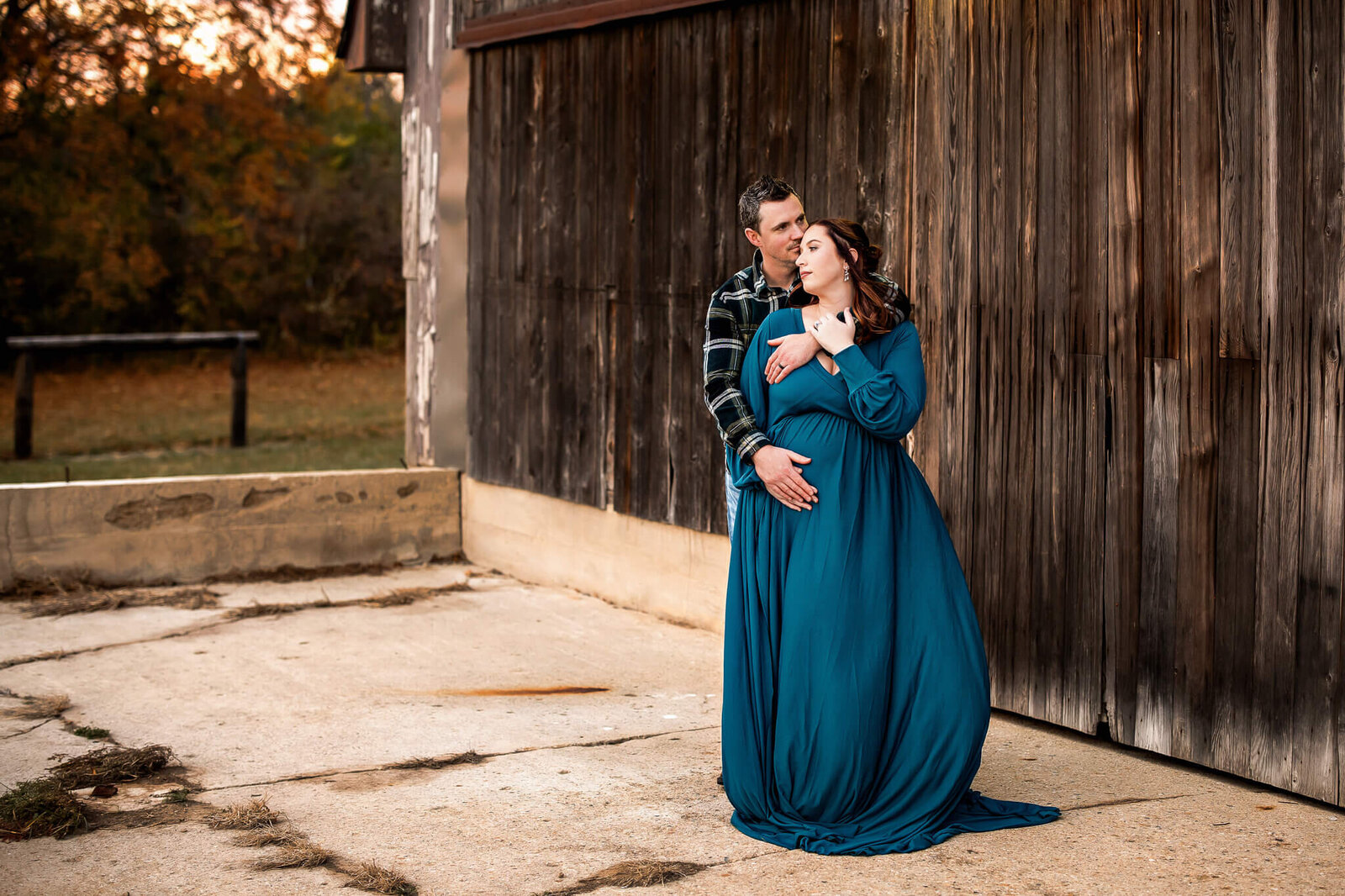 A couple stands outside by a rustic wooden wall as the man embraces the woman from behind, his hands on her pregnant belly. She wears a long teal dress; both look peaceful—perfect for maternity portraits with autumn trees in the background.